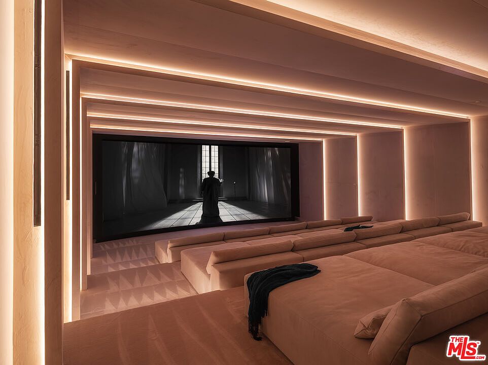 This image showcases a luxurious, minimalist home theater designed with tiered, plush seating and integrated warm LED cove lighting that accentuates the architectural depth of the room. The large, high-definition screen displays a cinematic scene, creating an immersive viewing experience within a sophisticated, monochromatic space. The perspective is from the back of the room, emphasizing the tiered layout and the cozy, lounge-like atmosphere.