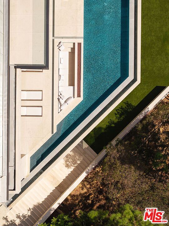 This striking aerial perspective captures a modern luxury estate, highlighting a sleek, geometric infinity-edge swimming pool that serves as the centerpiece of the outdoor living space. The design features a sunken lounge area adjacent to the water, clean stone decking, and a seamless transition into a lush, manicured lawn and surrounding natural landscape. The composition emphasizes architectural precision and a sophisticated, resort-like atmosphere perfect for high-end outdoor entertaining.