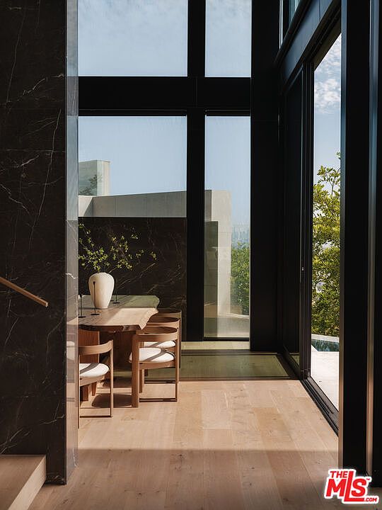 This sophisticated dining area features a live-edge wooden table paired with minimalist chairs, set against a dramatic dark marble accent wall. Floor-to-ceiling windows frame a serene outdoor view, bathing the light-toned hardwood floors in natural sunlight. The space exudes a modern, high-end aesthetic with a seamless blend of organic textures and architectural precision.