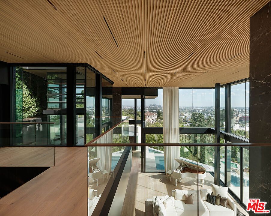 This stunning, double-height living room features a dramatic slatted wood ceiling and expansive floor-to-ceiling glass walls that frame a panoramic city view. The open-concept space includes a sleek white grand piano and a plush, modern seating area, all connected by a minimalist glass-railed mezzanine. The architectural design emphasizes luxury, light, and a seamless integration between the sophisticated interior and the outdoor landscape.