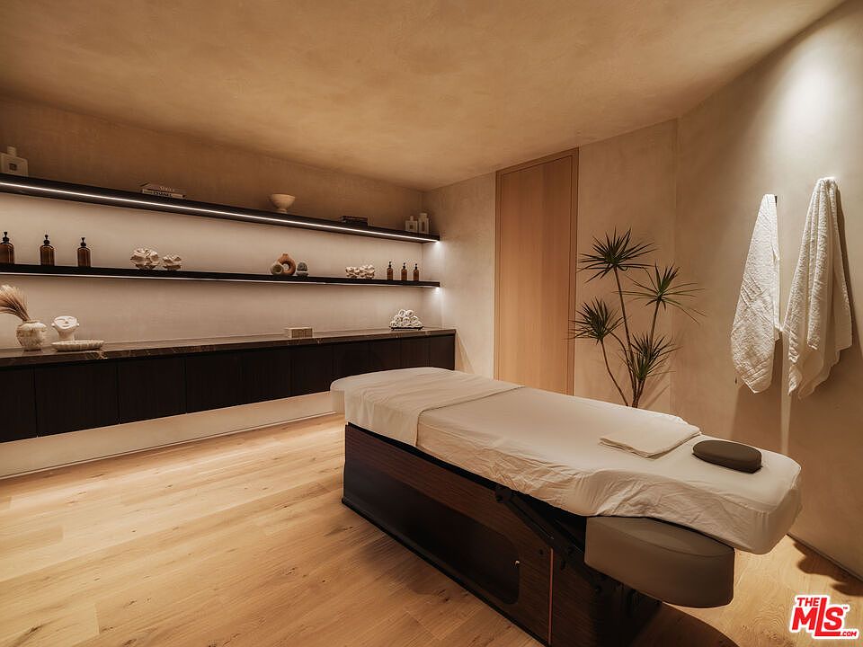 This serene spa or massage room features a minimalist, organic aesthetic with warm wood flooring and soft, neutral wall tones. A professional massage table sits center stage, complemented by sleek, illuminated floating shelves displaying curated decor and spa essentials. The space is designed for relaxation, with a clean, modern layout and a subtle, calming atmosphere.