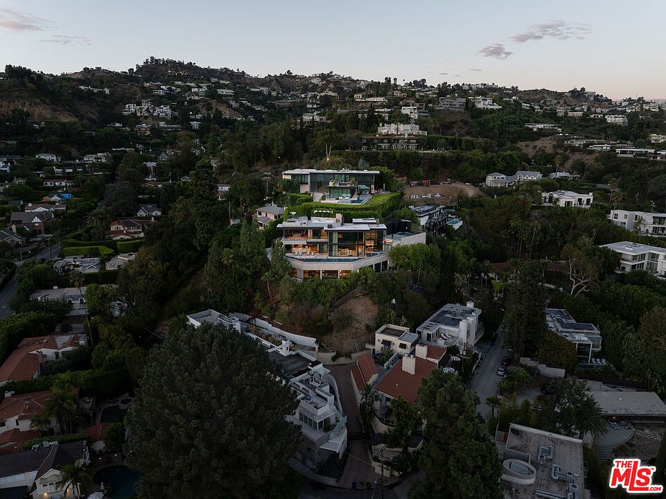 This high-angle aerial shot captures a sprawling, multi-level modern luxury estate nestled into a lush, hilly landscape in Los Angeles. The property features tiered architecture with expansive glass walls, multiple terraces, and a prominent swimming pool, all surrounded by dense greenery and neighboring residential homes. The perspective provides a cinematic overview of the home's integration into the hillside, emphasizing its secluded yet prominent position within the neighborhood.