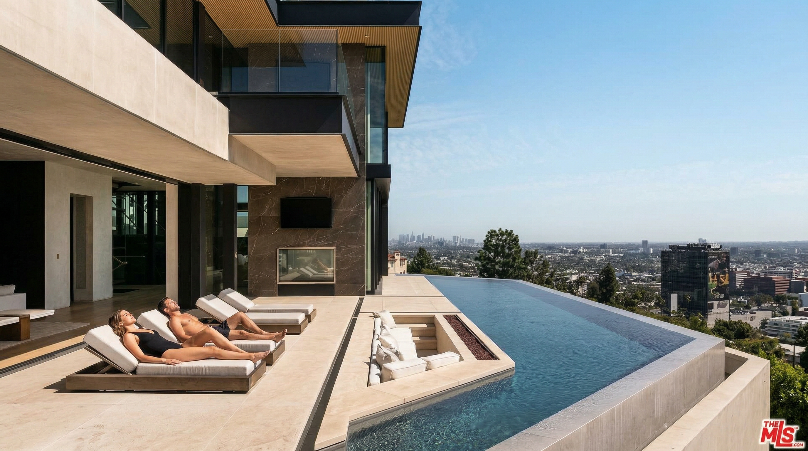 This stunning modern luxury estate features a seamless indoor-outdoor transition, highlighted by a sleek infinity-edge pool that overlooks a sprawling city skyline. The expansive stone deck includes a sunken lounge area with a fire pit and multiple chaise lounges, creating an ideal space for high-end entertaining. The architectural design emphasizes clean lines, dark stone accents, and floor-to-ceiling glass, offering a sophisticated and breathtaking panoramic view.