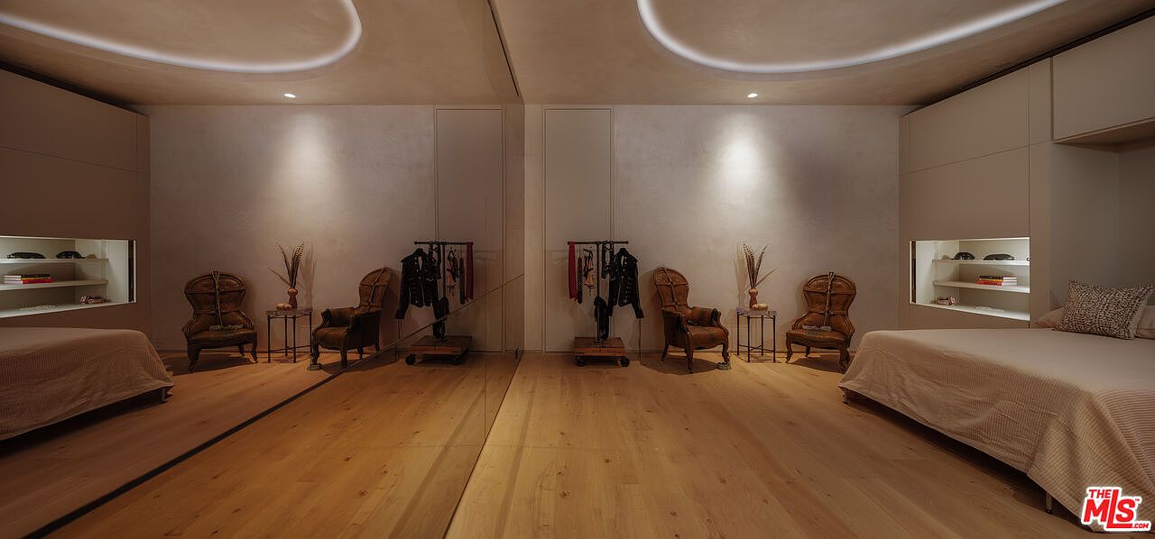 This sophisticated primary bedroom features a symmetrical layout with a large central mirror reflecting the room's elegant design. The space is anchored by two beds on either side, each accompanied by a vintage-style leather armchair and a small side table, all set against a backdrop of warm, light-toned wood flooring and recessed wall shelving. The minimalist aesthetic is enhanced by soft, curved ceiling lighting, creating a serene and luxurious atmosphere.