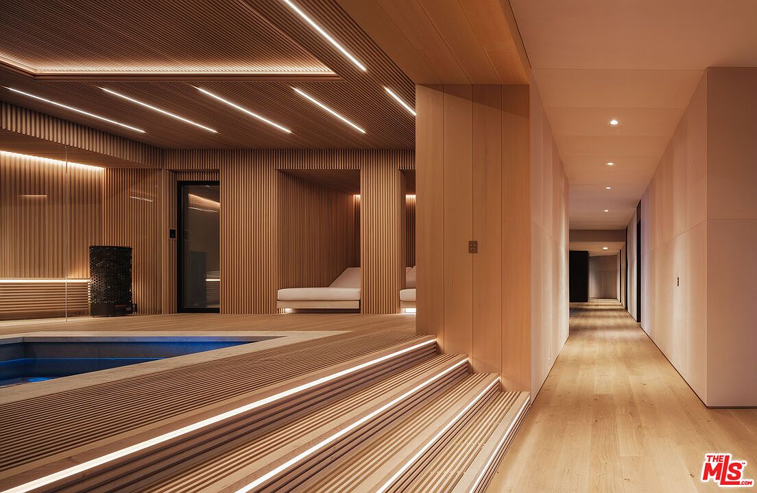 This sophisticated wellness area features a minimalist, spa-like aesthetic defined by extensive use of warm-toned, slatted wood paneling and integrated linear LED lighting. The space includes a sunken pool area with tiered steps leading to a serene lounge zone, seamlessly transitioning into a long, clean-lined hallway. The overall design exudes luxury and tranquility, emphasizing high-end craftsmanship and a cohesive, modern architectural style.