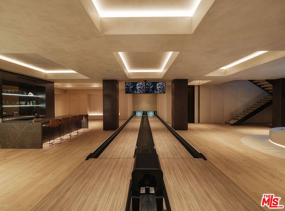 This image showcases a luxurious, modern home bowling alley featuring two lanes with polished wooden surfaces and a sleek, dark-toned bar area to the left. The space is illuminated by recessed cove lighting in the ceiling, creating a sophisticated and high-end atmosphere. A staircase with a glass railing is visible on the right, emphasizing the open-concept design and premium finishes of this entertainment space.