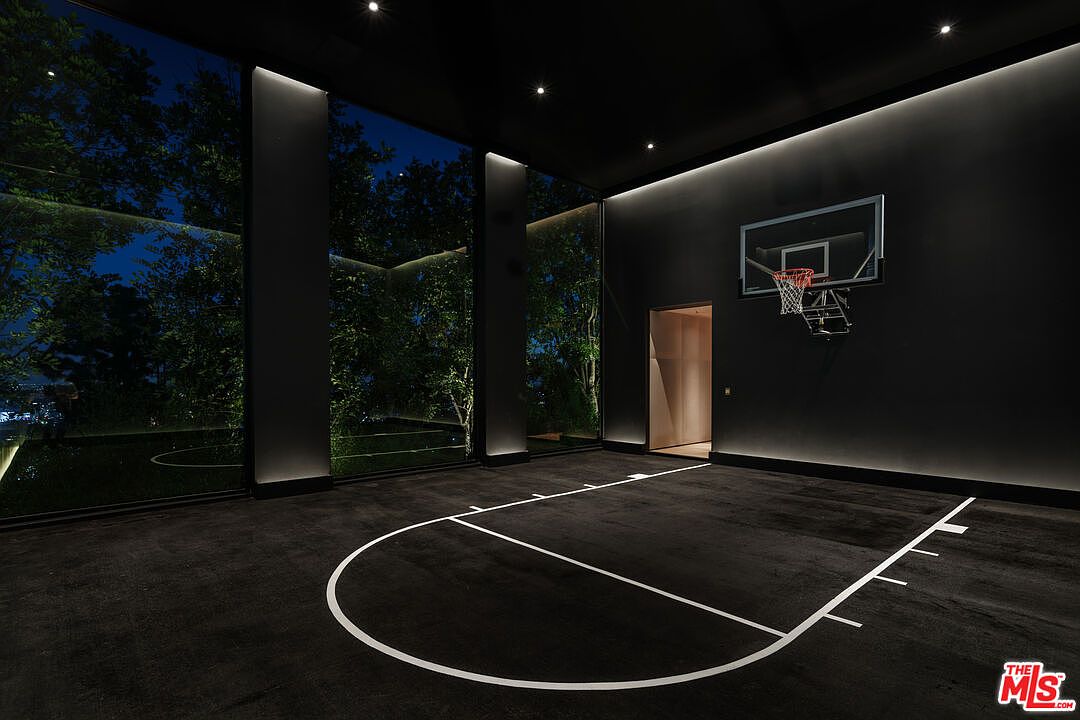 This sophisticated indoor basketball court features a sleek, monochromatic black aesthetic with floor-to-ceiling glass walls that offer a dramatic view of the illuminated night landscape. The space is minimalist and modern, highlighted by recessed ceiling lighting and a professional-grade basketball hoop mounted on a dark wall. The perspective captures the court's clean lines and luxurious, moody atmosphere, emphasizing its high-end design.