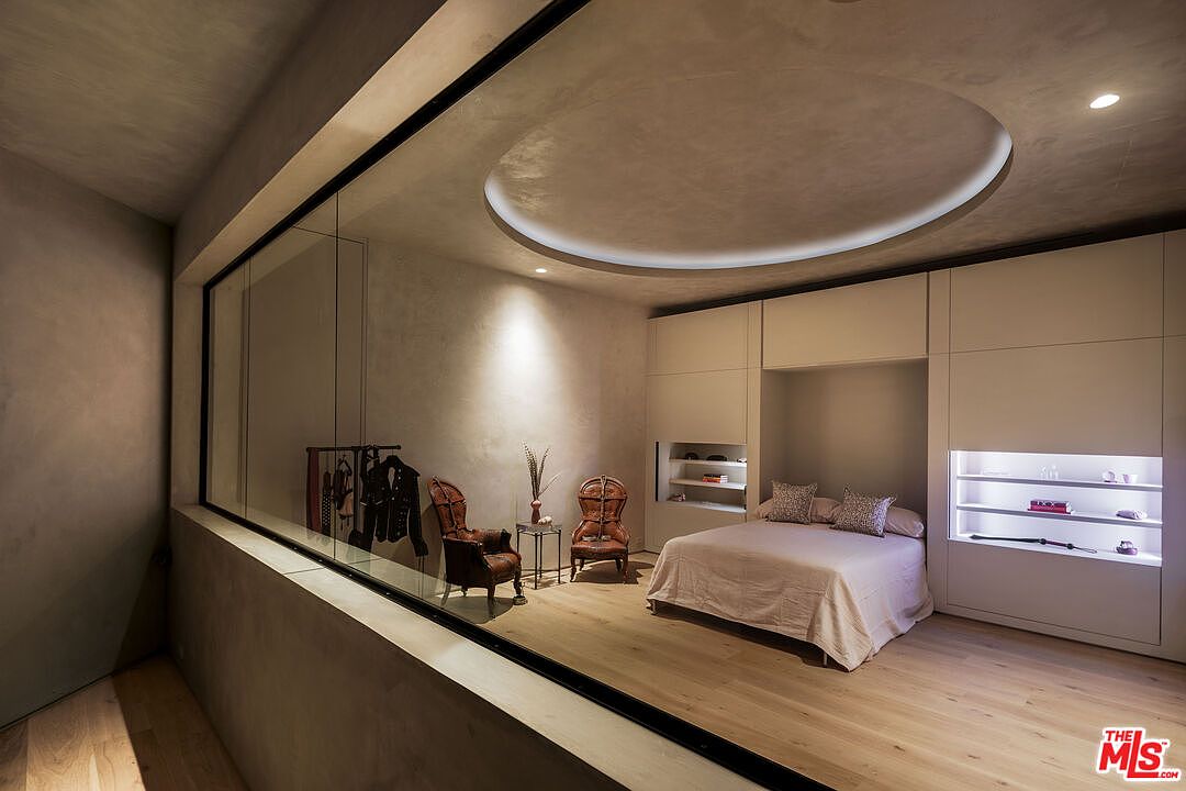 This sophisticated bedroom features a unique architectural design with a large glass partition separating the sleeping area from a lounge space. The room is characterized by a minimalist aesthetic, highlighted by a circular recessed ceiling light, light wood flooring, and custom built-in cabinetry with integrated shelving. The overall atmosphere is serene and modern, with a neutral color palette that emphasizes clean lines and high-end finishes.
