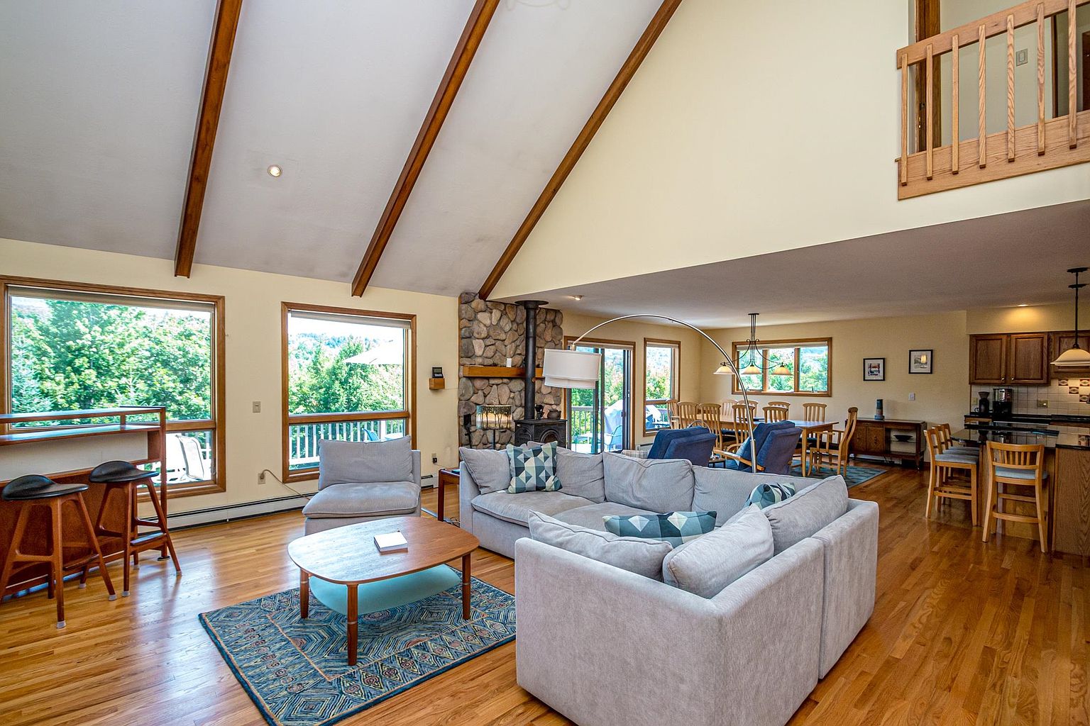 This spacious open-concept living area features a vaulted ceiling with exposed wooden beams and light-toned walls. The room is anchored by a large sectional sofa and a stone-clad chimney breast, while a breakfast bar and dining area are visible in the background. Large windows provide ample natural light and views of the surrounding greenery, creating an airy and inviting atmosphere.