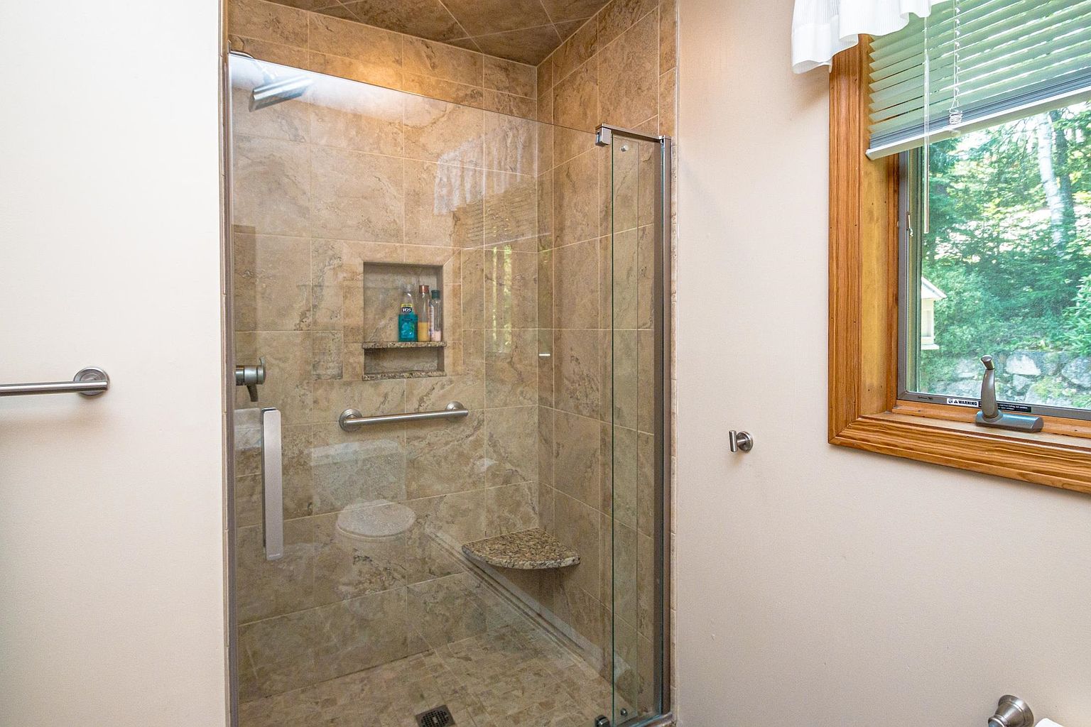 This bathroom features a walk-in shower enclosed by a glass door, showcasing neutral-toned stone tile walls and a built-in corner bench. A recessed niche provides convenient storage for toiletries, while a horizontal grab bar adds a functional safety element. To the right, a window with wooden trim allows natural light to brighten the space.