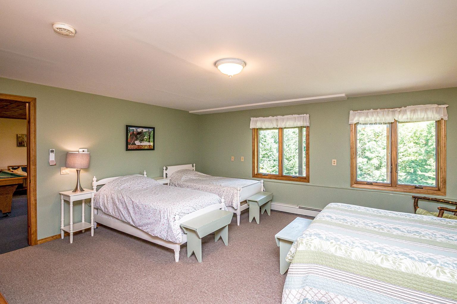 This spacious bedroom features a neutral color palette with sage green walls and light-toned carpeting. The room is furnished with two twin beds and a portion of a larger bed visible in the foreground, each accompanied by a simple wooden bench at the foot. Two large windows with white valances allow natural light to fill the space, while a doorway on the left provides a glimpse into an adjacent room with a pool table.