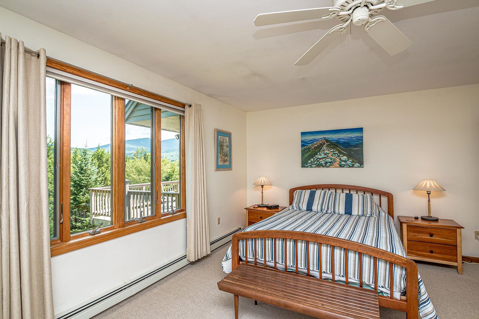 This cozy bedroom features a wooden bed frame with striped bedding and a matching wooden bench at the foot of the bed. Large windows with light-colored curtains offer a scenic view of the surrounding trees and mountains, while a ceiling fan and two bedside lamps provide lighting. The room is decorated with a landscape painting above the bed, creating a peaceful and inviting atmosphere.