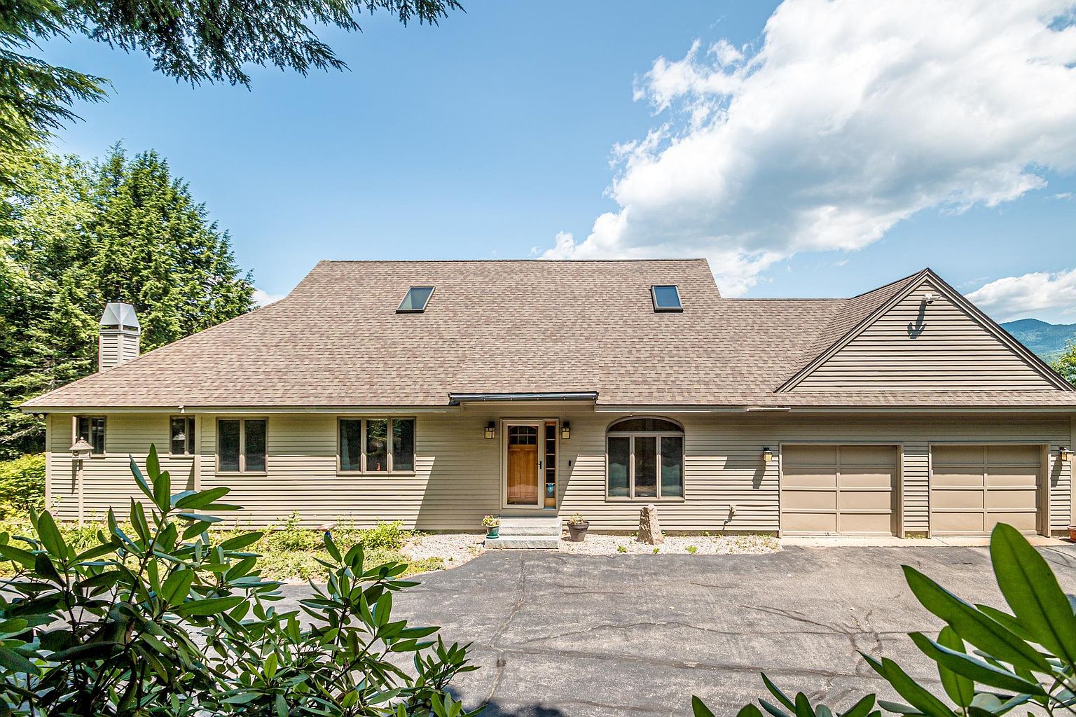 This front-facing view captures a suburban home with light-colored horizontal siding and a multi-gabled roofline featuring two skylights. The property includes an attached two-car garage, a central wooden front door, and a paved driveway leading up to the entrance. Lush green trees and shrubs frame the house, creating a welcoming and established residential atmosphere.
