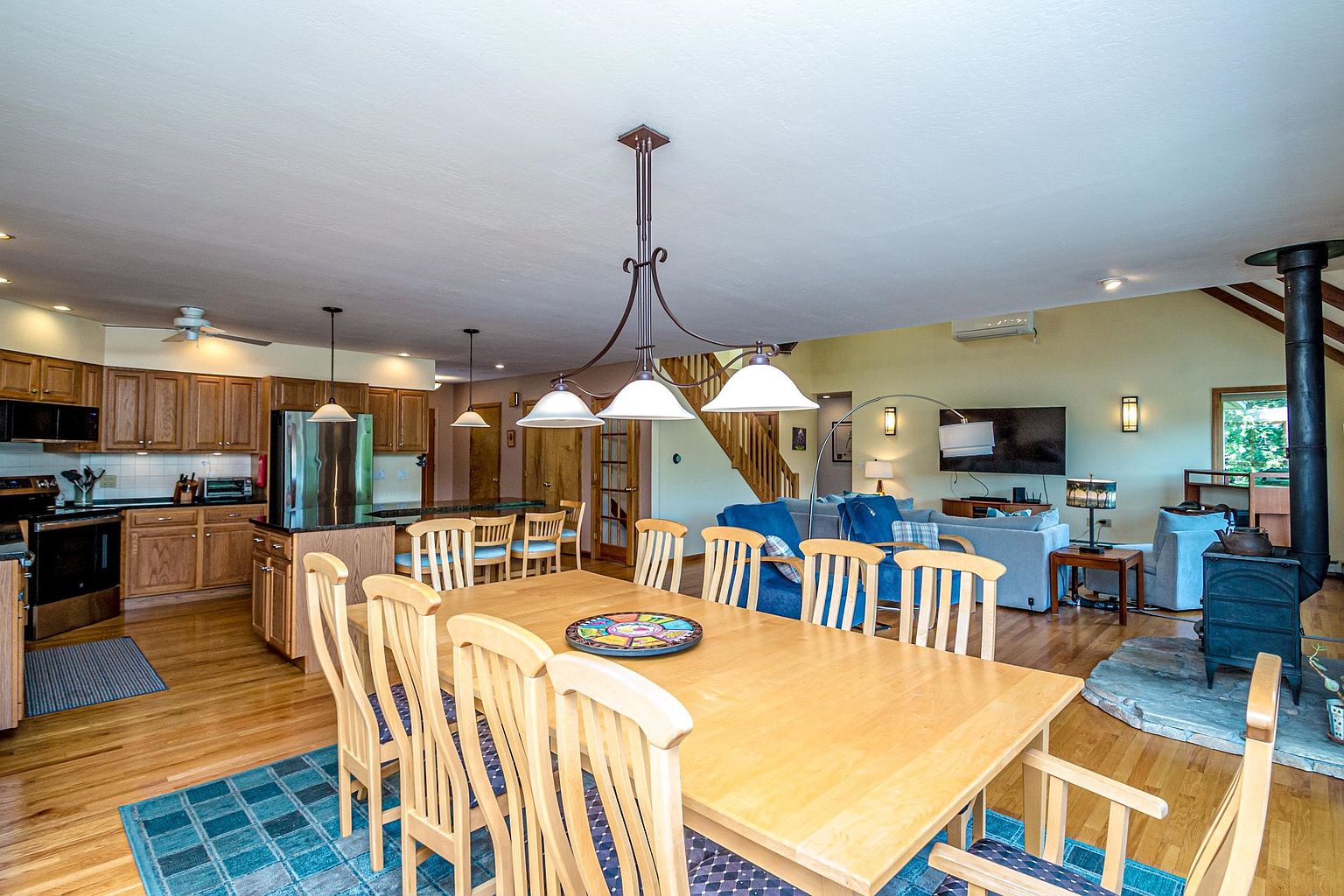 This open-concept living space features a prominent dining area in the foreground with a large light wood table and matching chairs set atop a blue patterned rug. The kitchen, visible to the left, includes light wood cabinetry and stainless steel appliances, while the living area in the background is anchored by a wood-burning stove on a stone hearth. The room is illuminated by a multi-light chandelier and recessed lighting, creating a warm and inviting atmosphere with a clear view of the staircase leading to the upper level.
