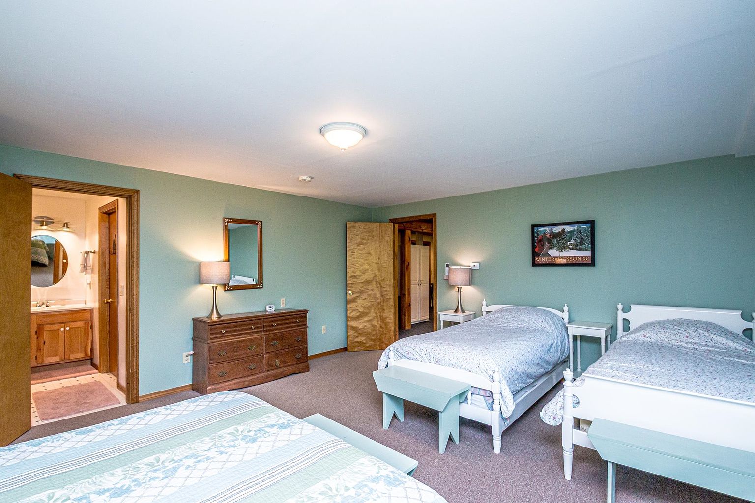 This spacious guest bedroom features two twin beds with light-colored bedding and matching wooden benches at the foot of each. The room includes a wooden dresser with a mirror, two bedside lamps, and a doorway leading to an en-suite bathroom. The walls are painted a soft sage green, creating a calm and inviting atmosphere.