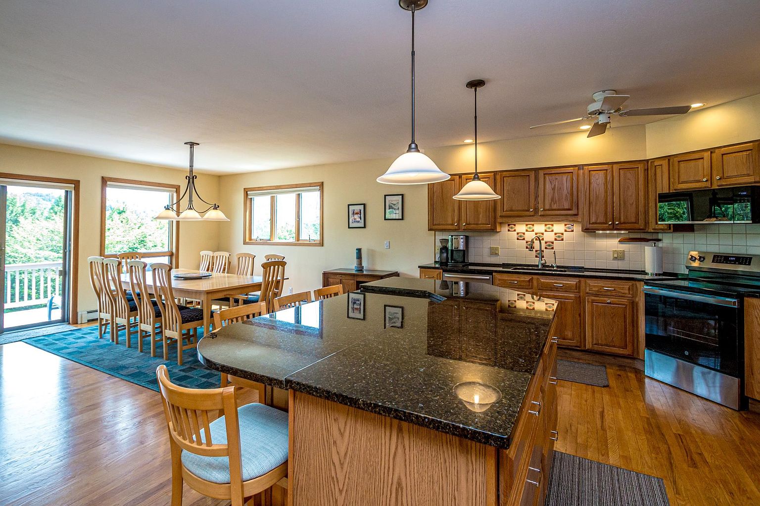 This open-concept kitchen and dining area features warm wood cabinetry, dark stone countertops, and a large central island with seating. The space is illuminated by pendant lighting over the island and a chandelier above the dining table, with natural light streaming in from the sliding glass doors and windows. The hardwood flooring flows throughout the room, creating a cohesive and inviting atmosphere suitable for both cooking and entertaining.