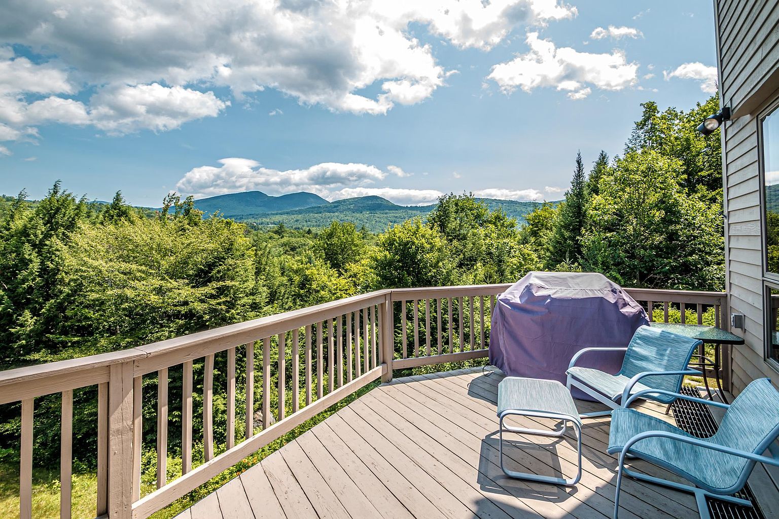 This elevated wooden deck offers a panoramic view of lush green mountains under a bright, partly cloudy sky. The space is furnished with two blue outdoor chairs and a matching footstool, alongside a covered grill, creating a peaceful spot for outdoor relaxation. The perspective captures the expansive scenery from the vantage point of the deck, highlighting the home's connection to the surrounding natural landscape.