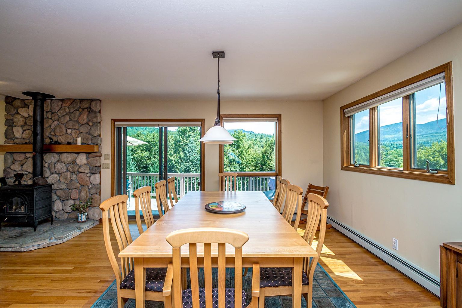 This dining room features a long light wood table surrounded by matching chairs with dark patterned cushions, set atop a blue area rug. To the left, a stone-clad chimney breast rises above a wood-burning stove, while large windows and a sliding glass door provide ample natural light and views of the surrounding trees and mountains. The space is illuminated by a single pendant light hanging above the table, creating a warm and inviting atmosphere.