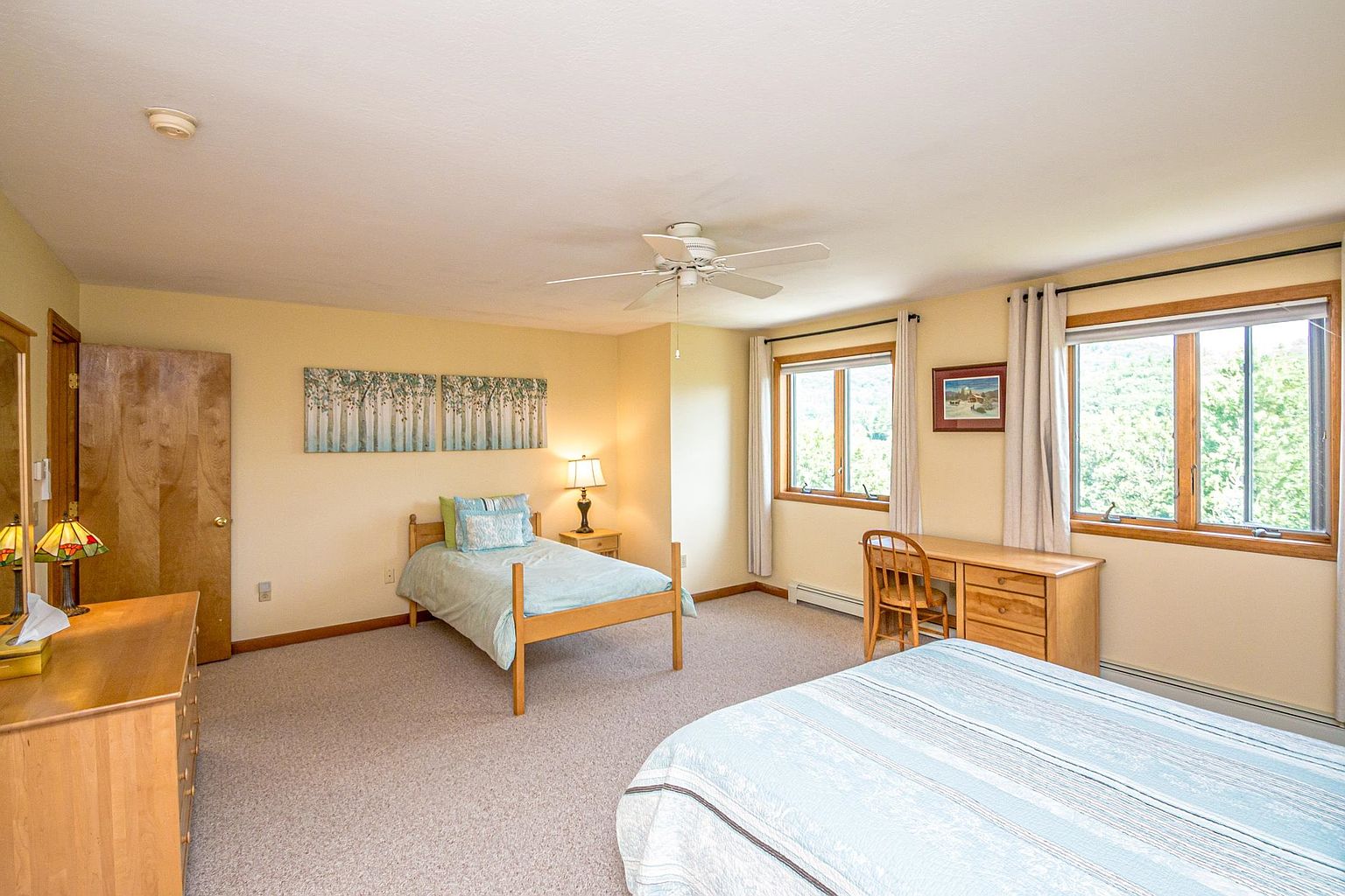 This spacious bedroom features a neutral color palette with light yellow walls and light-toned wood furniture, including a twin bed and a larger bed in the foreground. Two windows with light-colored curtains allow natural light to fill the room, which is accented by a piece of wall art depicting a forest scene. The room is carpeted and includes a wooden desk and chair, creating a functional and comfortable living space.