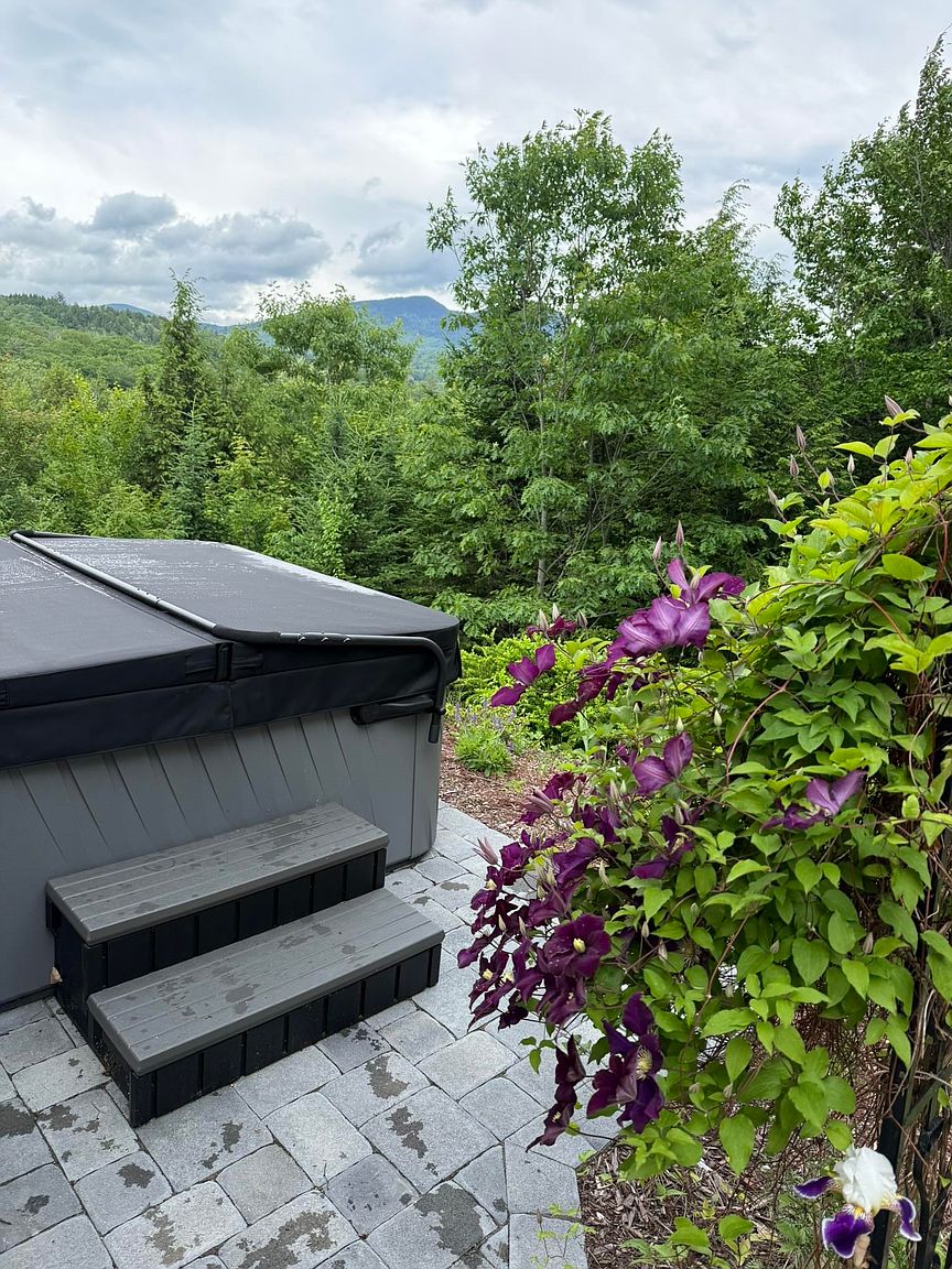 This outdoor space features a covered hot tub situated on a stone-paver patio, complete with a set of matching steps for easy access. To the right, a vibrant trellis is covered in blooming purple clematis flowers, adding a touch of natural beauty to the setting. The background showcases a scenic, elevated view of lush green trees and distant mountains under a cloudy sky, creating a serene and private atmosphere.
