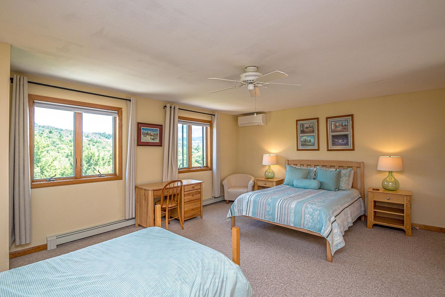This spacious guest bedroom features a light-filled layout with two beds, including a prominent bed with a light wood headboard and blue patterned bedding. The room is furnished with a matching light wood desk, nightstands, and a small armchair, all set against neutral walls and carpeted flooring. Large windows provide natural light and scenic views of the surrounding greenery, while a ceiling fan and wall-mounted climate control unit ensure comfort.