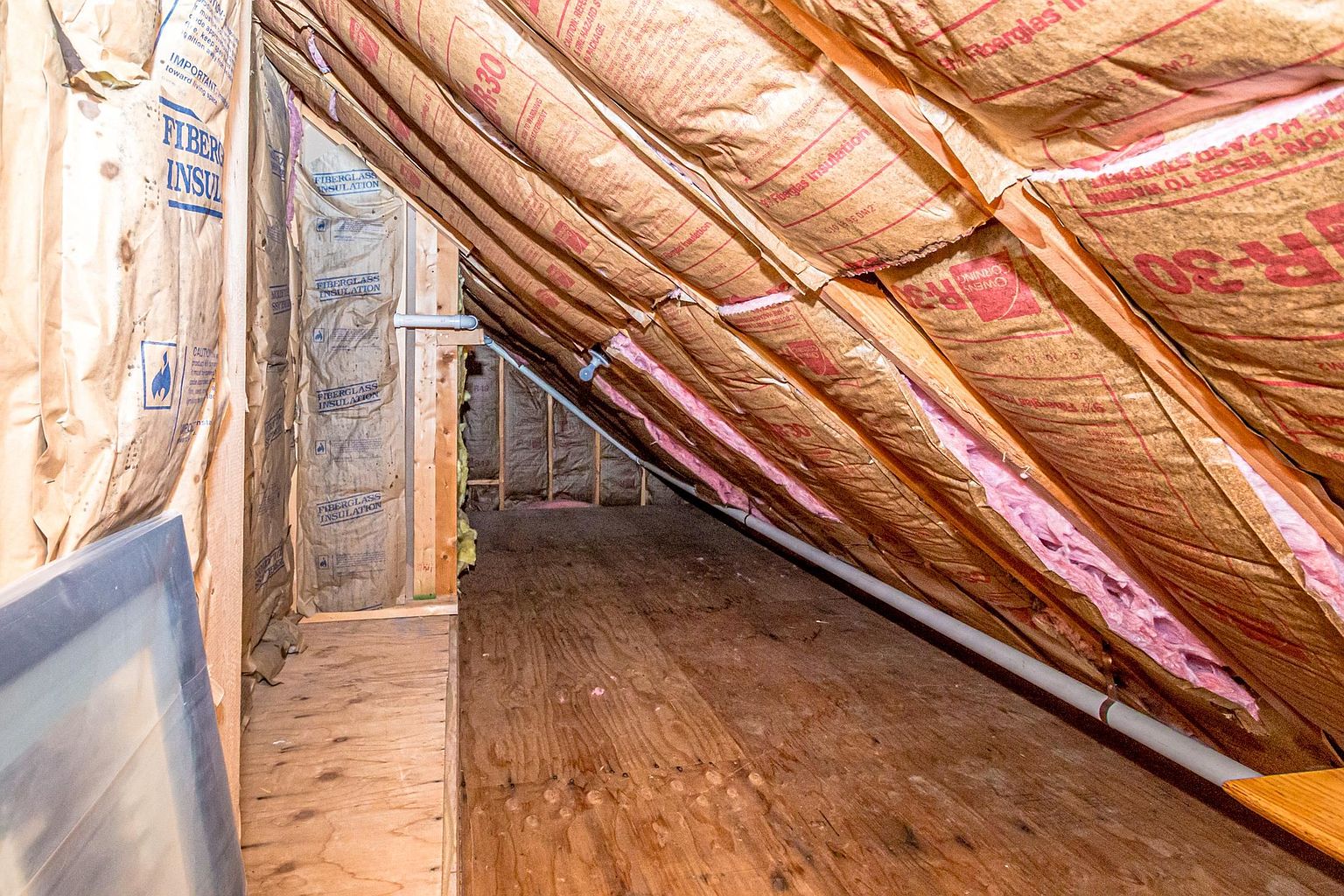 This image shows an unfinished attic space with exposed wooden framing and fiberglass insulation batts installed between the rafters. The perspective looks down a narrow, plywood-floored walkway that extends into the dark, sloped eaves of the roof structure. The scene highlights the functional, utilitarian nature of the home's upper storage or utility area.