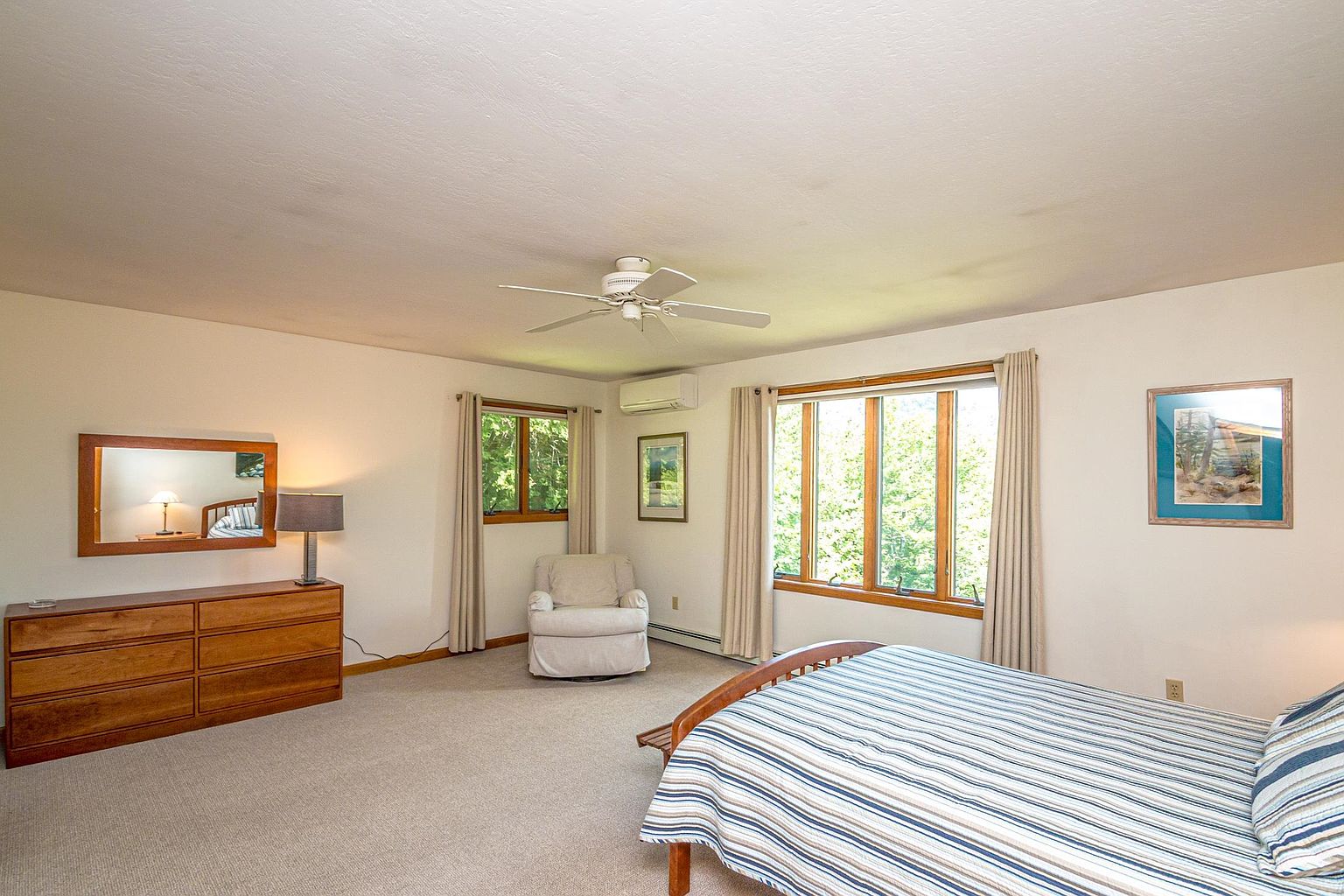 This spacious bedroom features neutral walls, light-toned carpeting, and a large wooden bed frame with a striped comforter. A wooden dresser with a mirror sits against the left wall, complemented by a cozy armchair tucked near a window. The room is brightened by natural light streaming through multiple windows, creating a calm and inviting atmosphere.