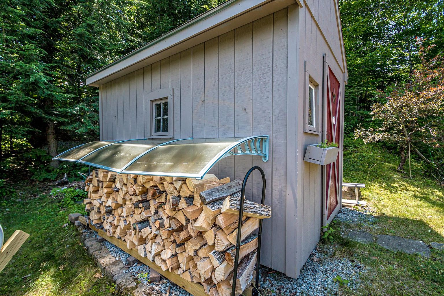 This side view of a small, light-colored wooden shed highlights a practical outdoor storage setup, featuring a neatly stacked woodpile protected by a modern, translucent curved awning. The structure includes a small window and a side door with a decorative star accent and a mounted planter box. The shed is situated in a lush, wooded setting with a gravel base, presenting a tidy and functional backyard utility area.