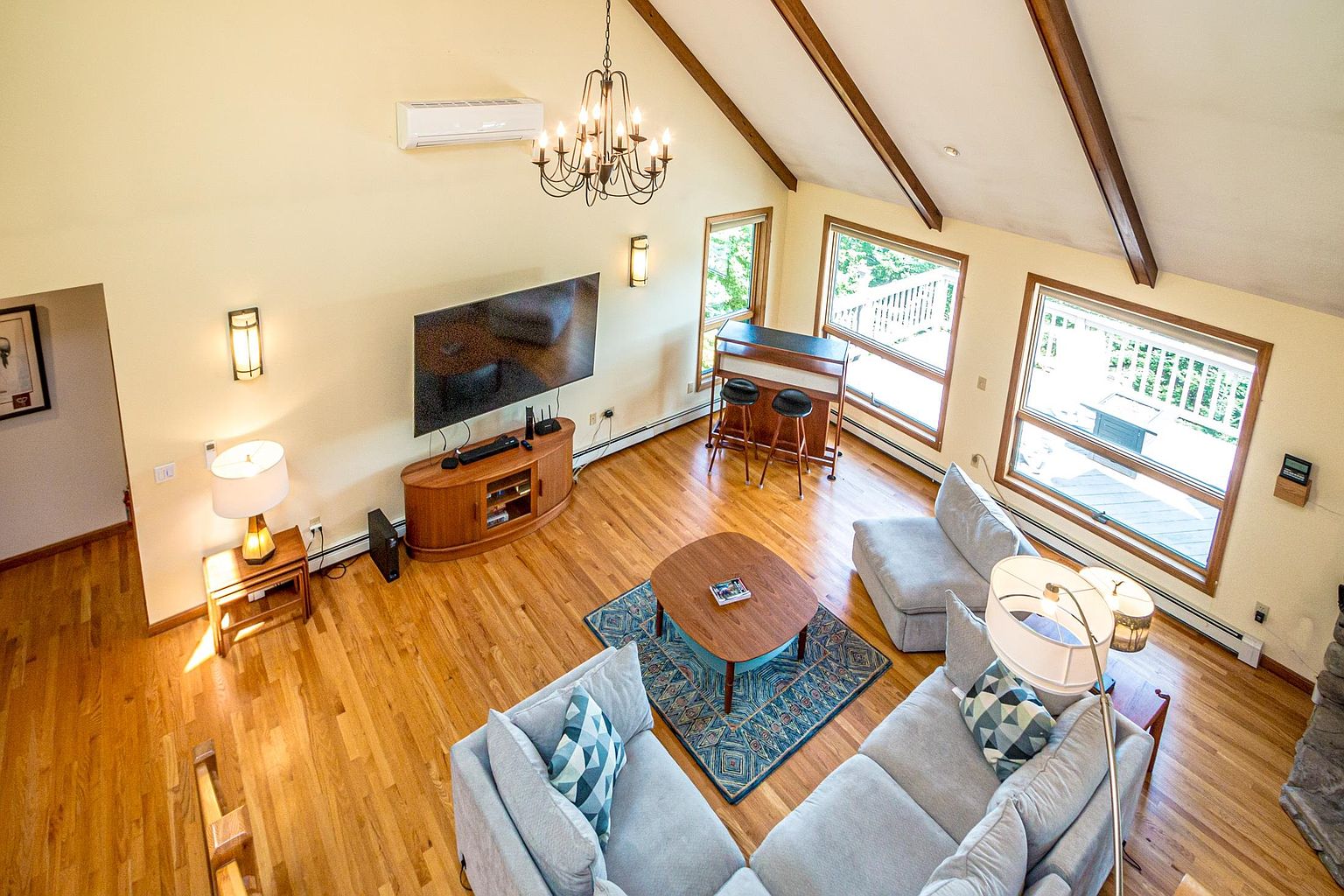 This high-angle shot captures a spacious, open-concept living room with vaulted ceilings and exposed wooden beams. The room features light-toned hardwood flooring, a large television mounted above a wooden media console, and a comfortable seating area arranged around a central coffee table on a patterned rug. Large windows provide ample natural light, while a small bar area with stools adds a functional touch to the airy, inviting space.