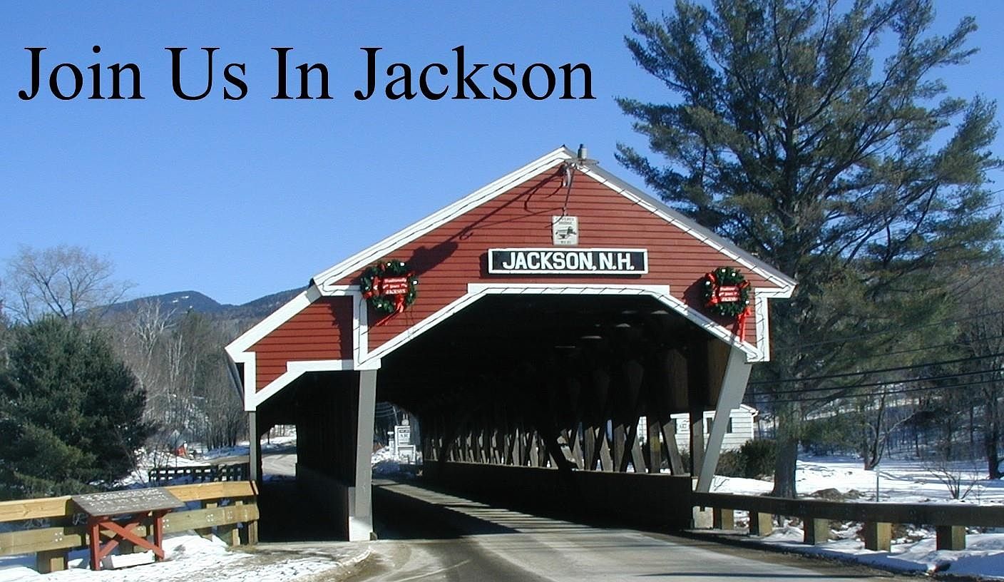 This image features the iconic red covered bridge in Jackson, New Hampshire, set against a clear blue sky and a snowy landscape. The structure is framed by a large, bare tree on the right and distant mountains in the background, creating a picturesque, seasonal scene. The bridge is adorned with festive wreaths, emphasizing its role as a charming local landmark.
