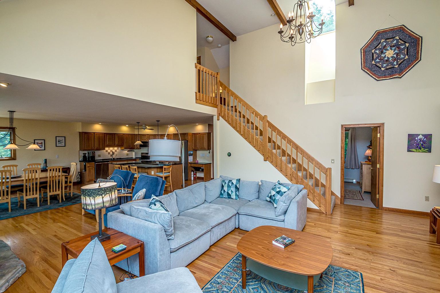 This open-concept living area features a spacious sectional sofa arranged around a wooden coffee table, set against a backdrop of light-toned walls and hardwood flooring. A wooden staircase with a matching railing leads to an upper level, while a large chandelier hangs from the vaulted ceiling above. The space flows seamlessly into a kitchen area in the background, creating an airy and inviting atmosphere suitable for family gatherings.