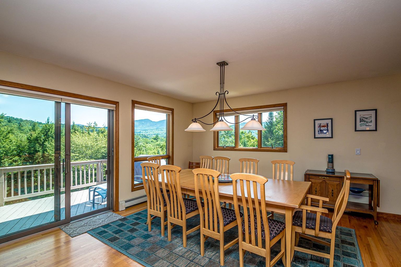 This bright dining room features a light wood table surrounded by six matching chairs, all set atop a blue and green patterned area rug. A three-light chandelier hangs above the table, while large sliding glass doors provide access to an outdoor deck and offer expansive mountain views. The room is finished with neutral walls, hardwood flooring, and a wooden sideboard against the wall adorned with framed artwork.