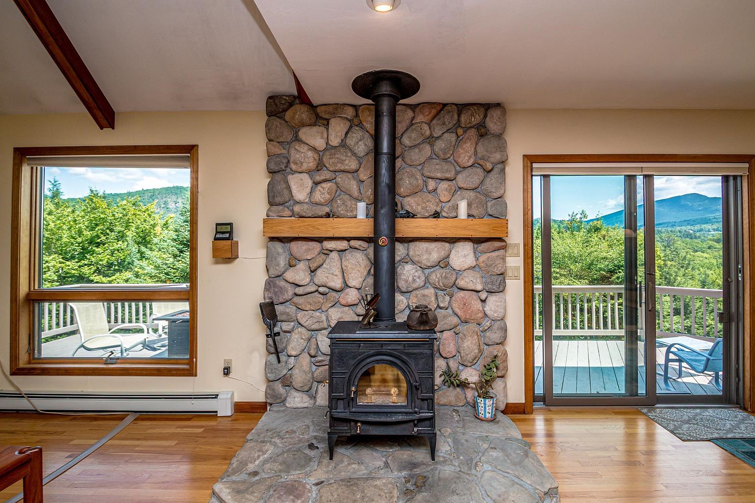 This living room features a prominent wood-burning stove set upon a raised stone hearth, centered against a stone-clad chimney breast. A horizontal wood mantel spans the stone wall, while large windows and a sliding glass door on either side provide abundant natural light and views of the surrounding forest. The space is finished with light wood flooring, creating a warm and rustic atmosphere.