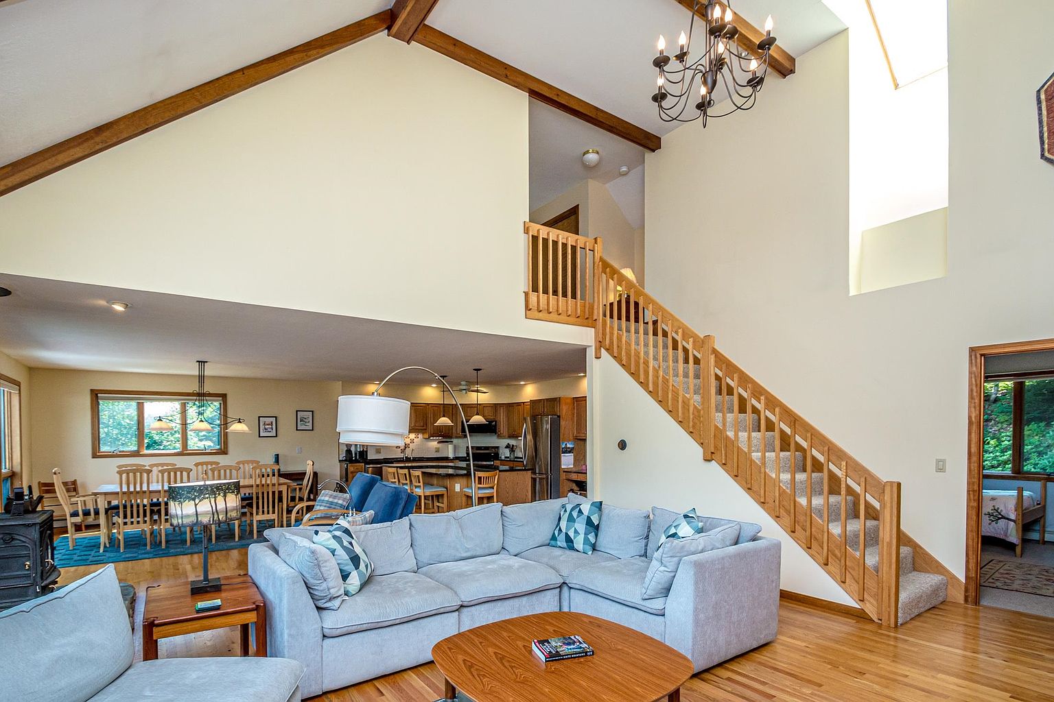 This open-concept living area features a vaulted ceiling with exposed wooden beams and a large chandelier, creating an airy and spacious atmosphere. A comfortable light-colored sectional sofa faces a wooden coffee table, while a staircase with wooden railings leads to an upper level. In the background, a dining area and a kitchen with wooden cabinetry are visible, showcasing a cohesive and functional layout.