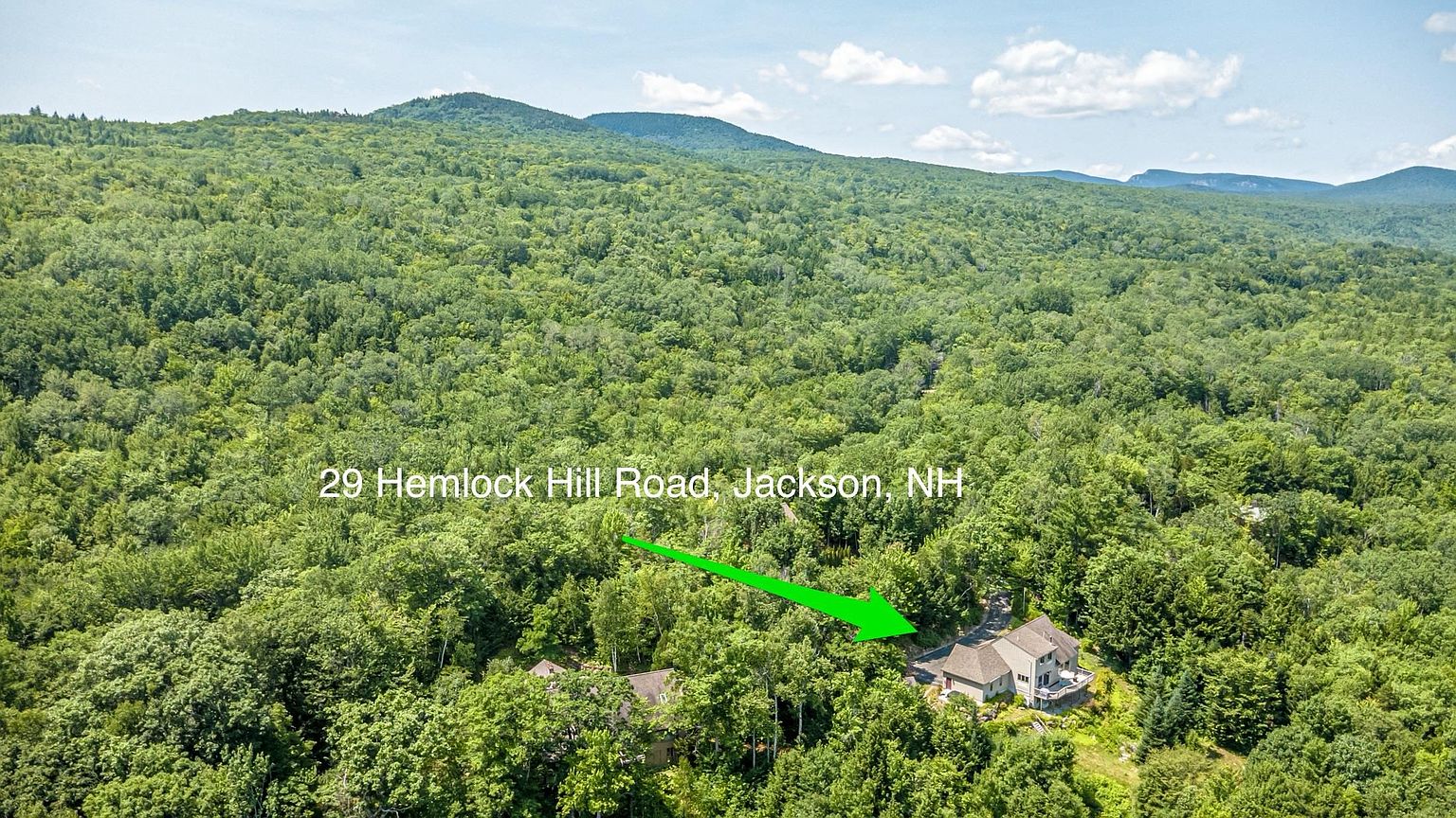 This high-angle aerial shot captures a multi-level residential home nestled within a dense, lush forest in Jackson, NH. The property features a multi-pitched roofline and a prominent deck overlooking the surrounding woodland, with a winding driveway leading to the structure. The expansive green canopy of the mountains provides a serene and secluded backdrop for the home.
