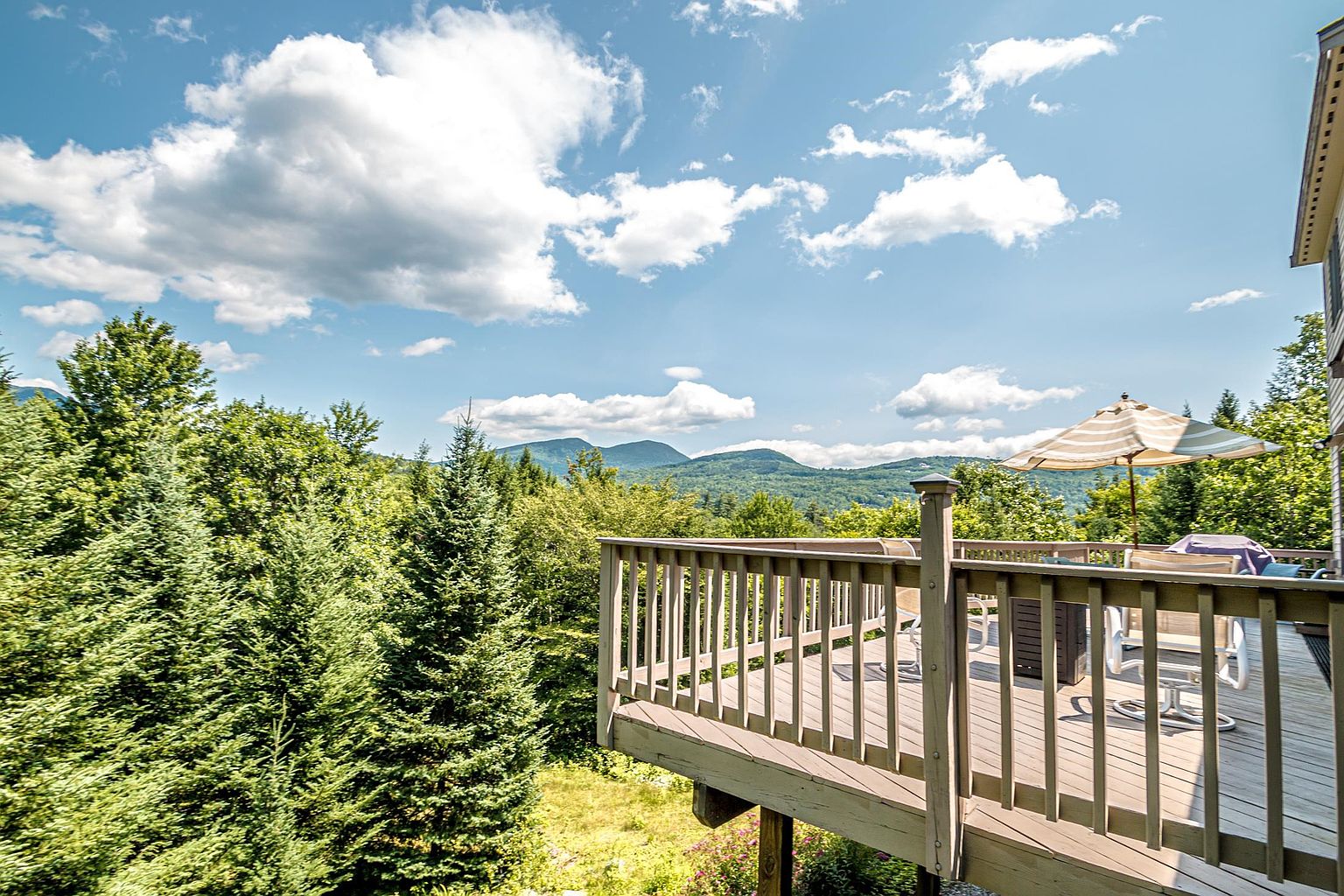This elevated wooden deck offers a panoramic view of lush green mountains under a bright blue sky with scattered clouds. The deck is furnished with outdoor seating and a striped umbrella, providing a perfect vantage point for enjoying the surrounding forest landscape. The perspective captures the expansive outdoor living space as it transitions from the home into the natural scenery.