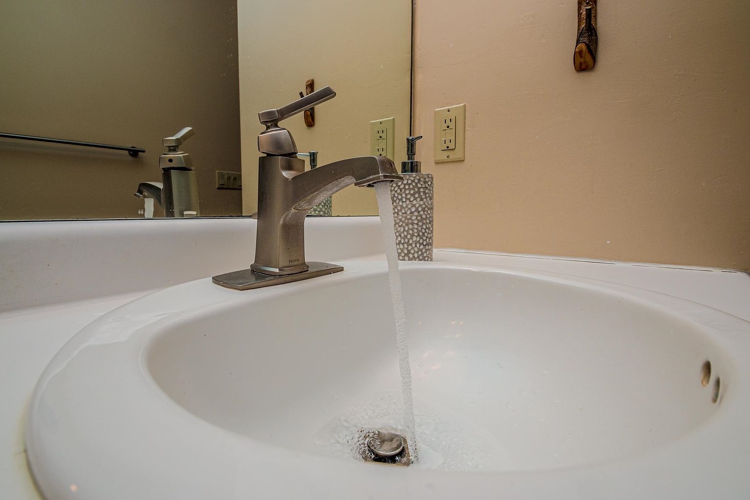 A close-up, eye-level shot of a bathroom vanity sink with a brushed-nickel faucet actively running water into the basin. The scene includes a textured, light-colored soap dispenser and a mirror reflecting the faucet and a wall-mounted electrical outlet. The neutral-toned wall and clean white sink create a functional and well-maintained appearance.