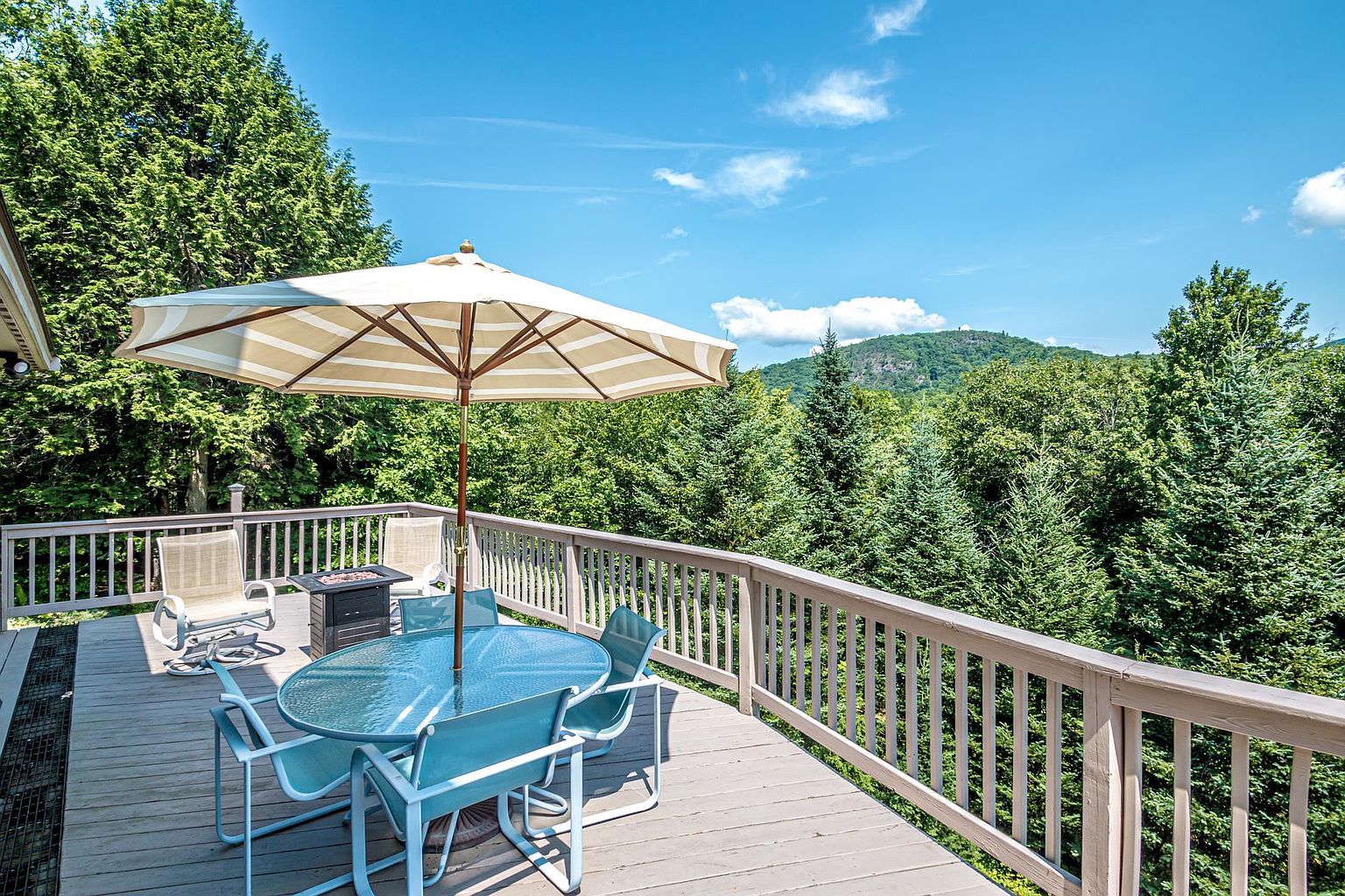 This elevated wooden deck offers a serene outdoor living space, complete with a glass-topped dining table shaded by a large, striped umbrella. The deck is enclosed by a simple wooden railing and overlooks a lush, dense forest with a mountain peak visible in the distance under a bright blue sky. The perspective captures the inviting atmosphere of the space, highlighting its potential for relaxation and scenic dining.