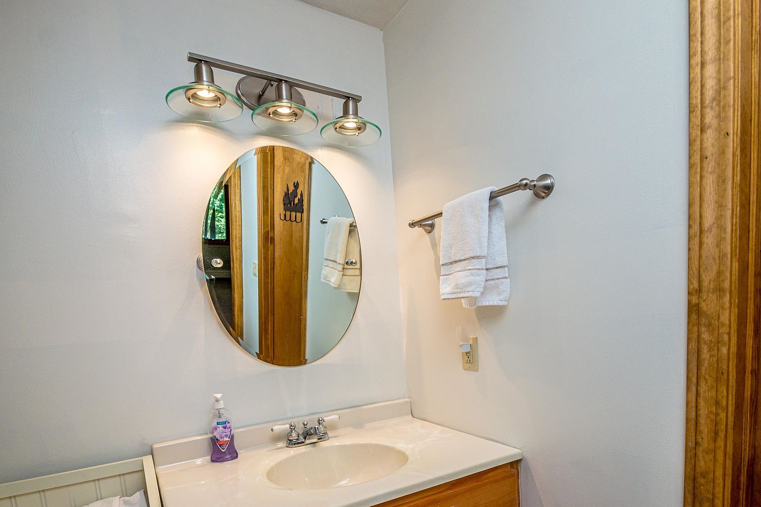 This bathroom vanity area features a light-toned countertop with an integrated sink and a simple faucet fixture. Above the vanity, a three-light vanity fixture illuminates an oval mirror, which reflects a glimpse of an adjacent room. A towel bar mounted on the wall holds a white towel, completing this functional and compact bathroom space.