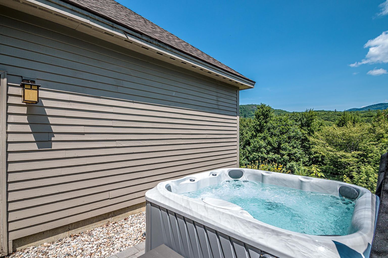 This exterior view highlights a modern hot tub situated on a paved patio area adjacent to the side of a house with light-colored horizontal siding. The spa is filled with bubbling water and features integrated headrests, set against a backdrop of lush green trees and a clear blue sky. The perspective captures the relaxing outdoor amenity, emphasizing the private and scenic setting of the property.