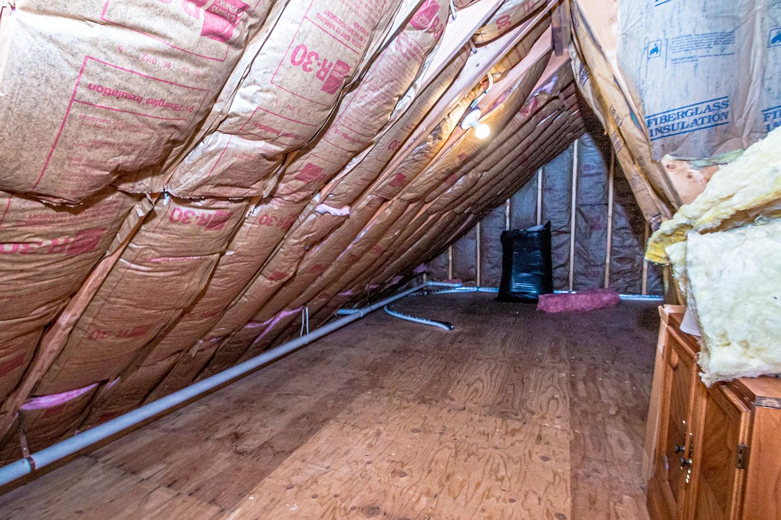 This attic space features sloped ceilings insulated with pink fiberglass batts, creating a cozy, enclosed atmosphere. The floor is covered in plywood sheeting, and a single light bulb provides illumination, highlighting the raw, functional nature of the area. A white pipe runs along the lower edge of the slope, and a black storage bag sits in the background, suggesting the space is primarily used for storage.