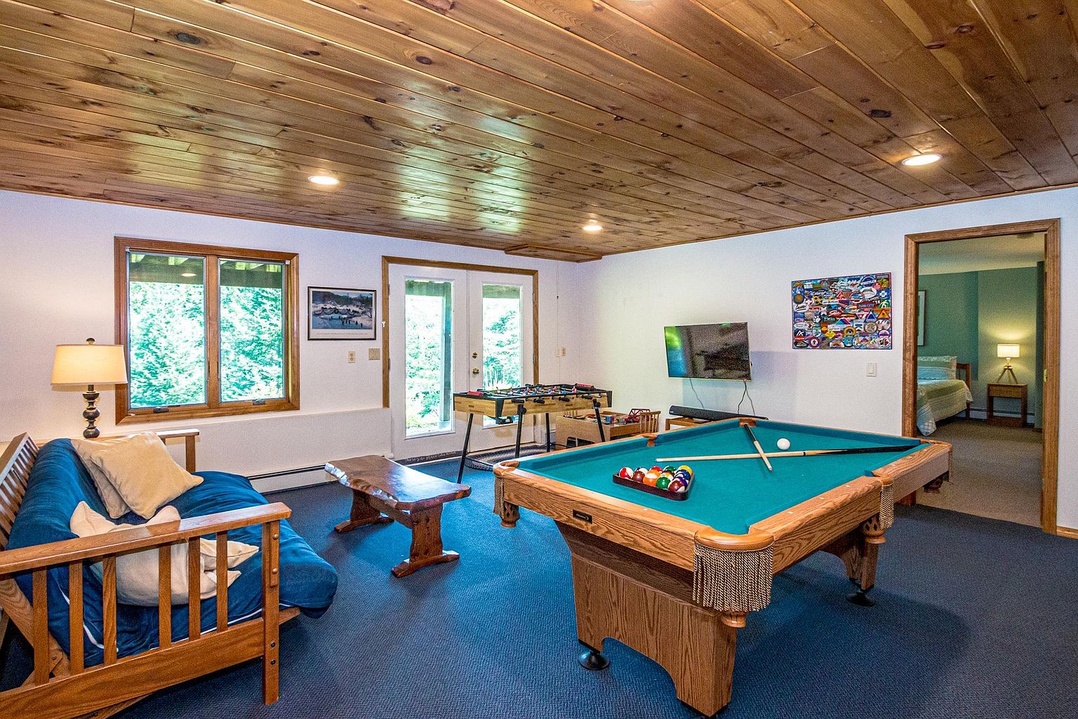 This spacious recreation room features a pool table as the central focal point, complemented by a foosball table and a blue futon seating area. The room is finished with a warm wood-paneled ceiling and blue carpeting, creating a casual and inviting atmosphere for entertainment. A doorway leads into an adjacent bedroom, while large windows and glass doors provide natural light and a view of the outdoors.