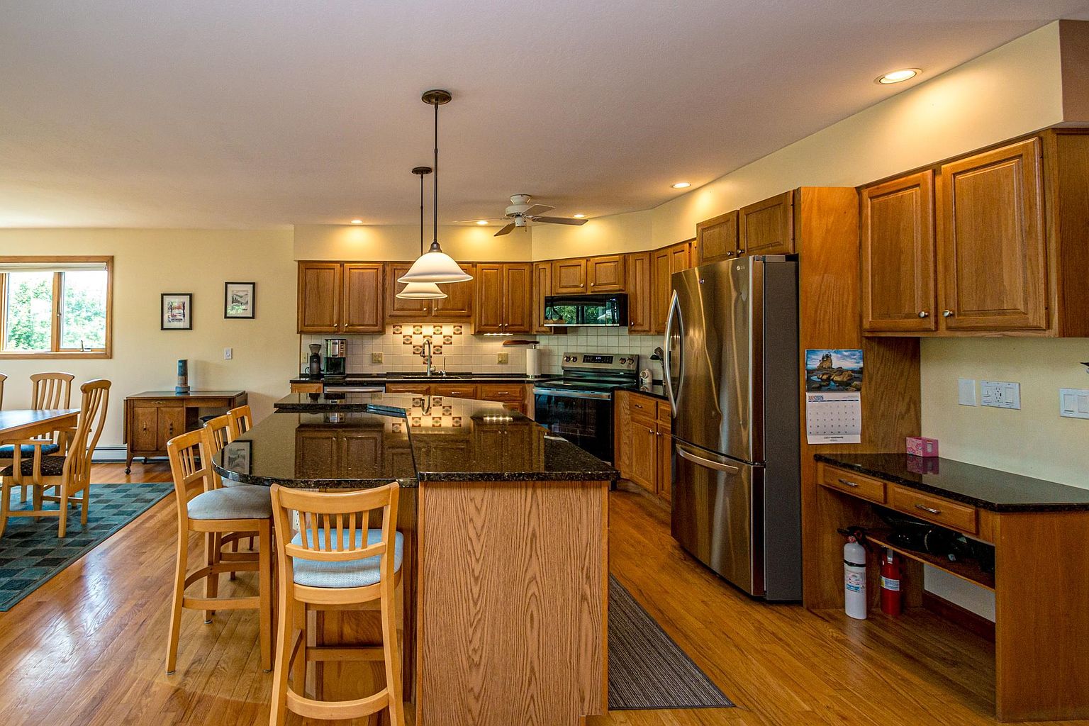 This spacious kitchen features warm-toned wood cabinetry, dark stone countertops, and a large central island with bar seating. The layout includes stainless steel appliances, a tiled backsplash, and pendant lighting, creating a functional and inviting cooking space. The open-concept design flows seamlessly into an adjacent dining area, with hardwood flooring throughout.