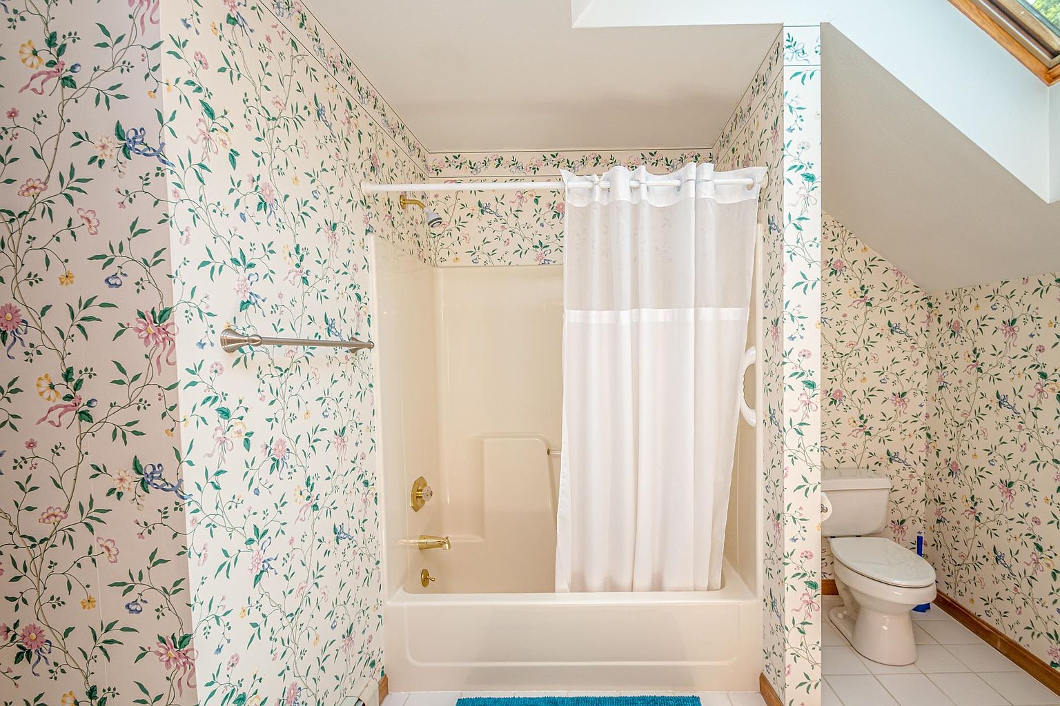 This bathroom features a classic tub-shower combination with a white curtain and a beige surround. The walls are covered in a traditional floral wallpaper, and the space includes a standard white toilet and white tile flooring. A skylight is visible in the upper right corner, providing natural light to the room.