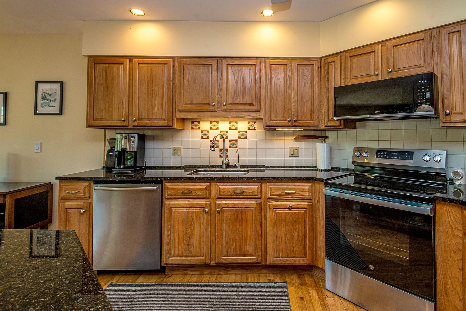 This kitchen features warm-toned wood cabinetry paired with dark stone countertops and a stainless steel appliance suite, including a dishwasher, microwave, and range. A decorative tile backsplash with a geometric pattern adds a focal point above the sink, while recessed lighting provides bright illumination across the workspace. The room is finished with hardwood flooring and a striped runner rug, creating a functional and inviting culinary space.