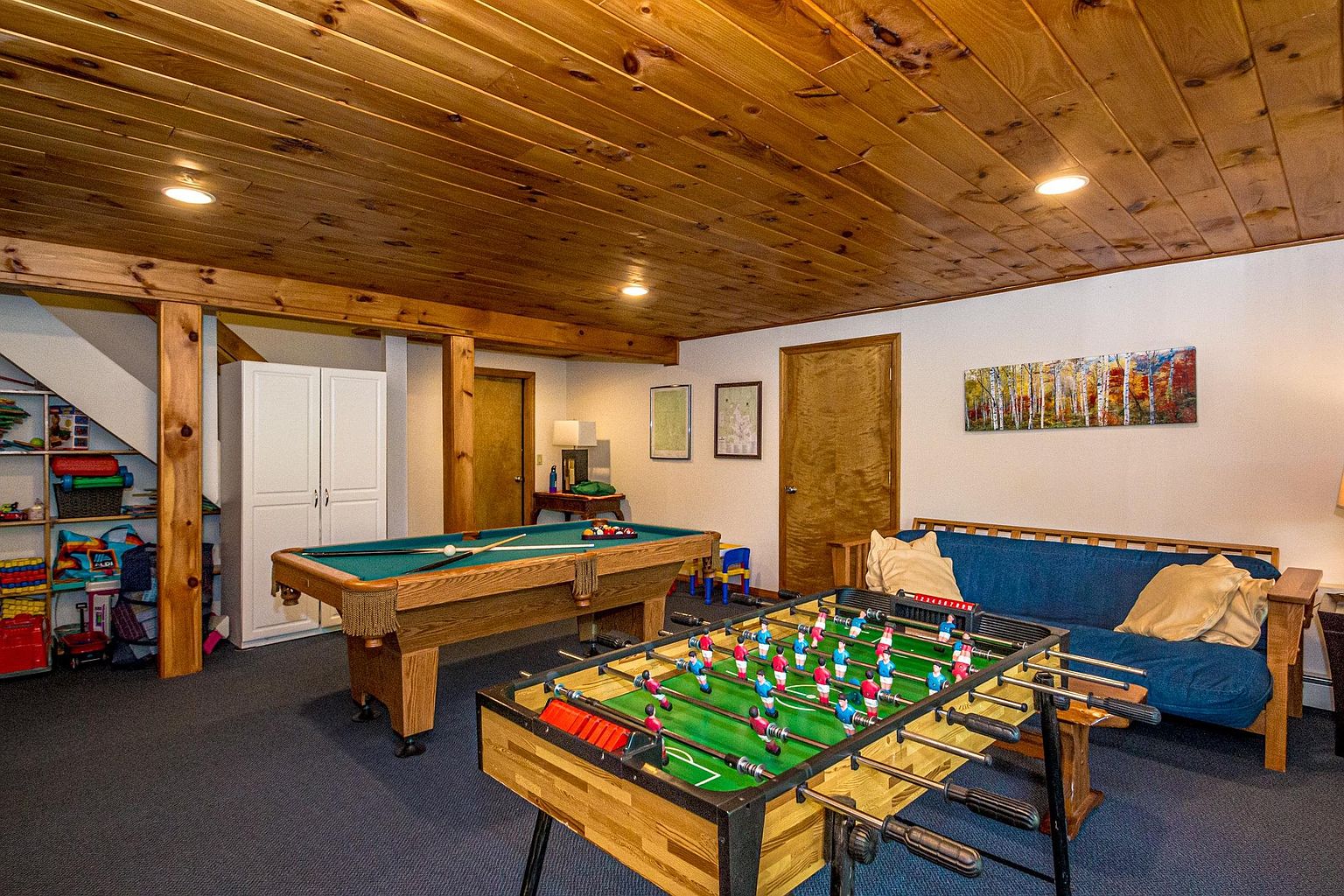 This spacious basement recreation room features a wood-paneled ceiling and dark carpeting, creating a cozy atmosphere for entertainment. The room is furnished with a pool table, a foosball table, and a blue futon, making it an ideal space for leisure activities. A white storage cabinet and open shelving under the stairs provide practical storage solutions, while a landscape painting adds a decorative touch to the wall.