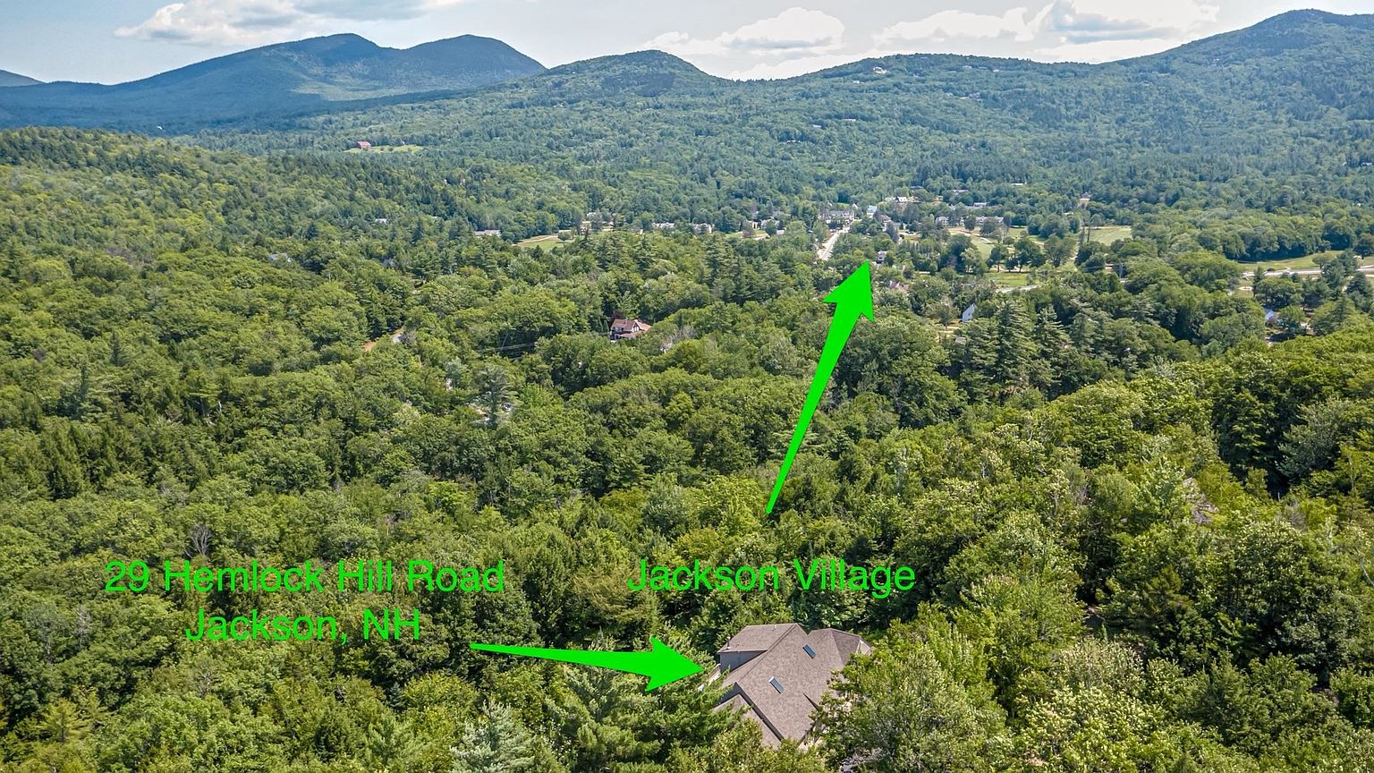 This aerial perspective captures a residential property nestled within a dense, lush forest, highlighting its secluded setting. The view showcases the home's roofline and its proximity to the nearby Jackson Village, which is indicated by a green arrow. The surrounding landscape features rolling hills and extensive greenery, emphasizing the peaceful and scenic nature of the location.