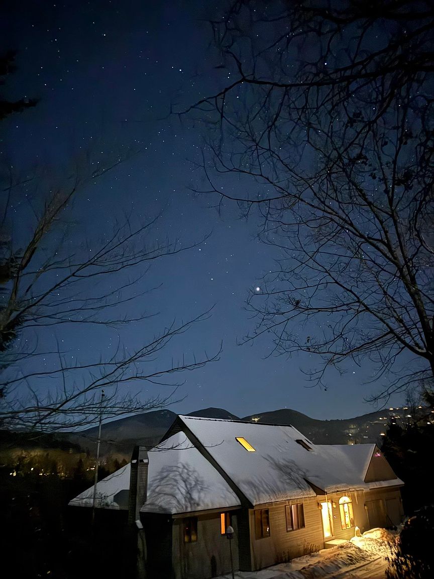 This evening exterior shot captures a cozy, multi-level home nestled against a backdrop of snow-dusted mountains under a clear, star-filled sky. The house features a prominent roofline with a skylight glowing from within, while warm interior lighting spills from the windows and entryway, creating an inviting contrast against the cool blue twilight. Bare tree branches frame the composition, adding depth and a sense of seasonal tranquility to this picturesque mountain residence.
