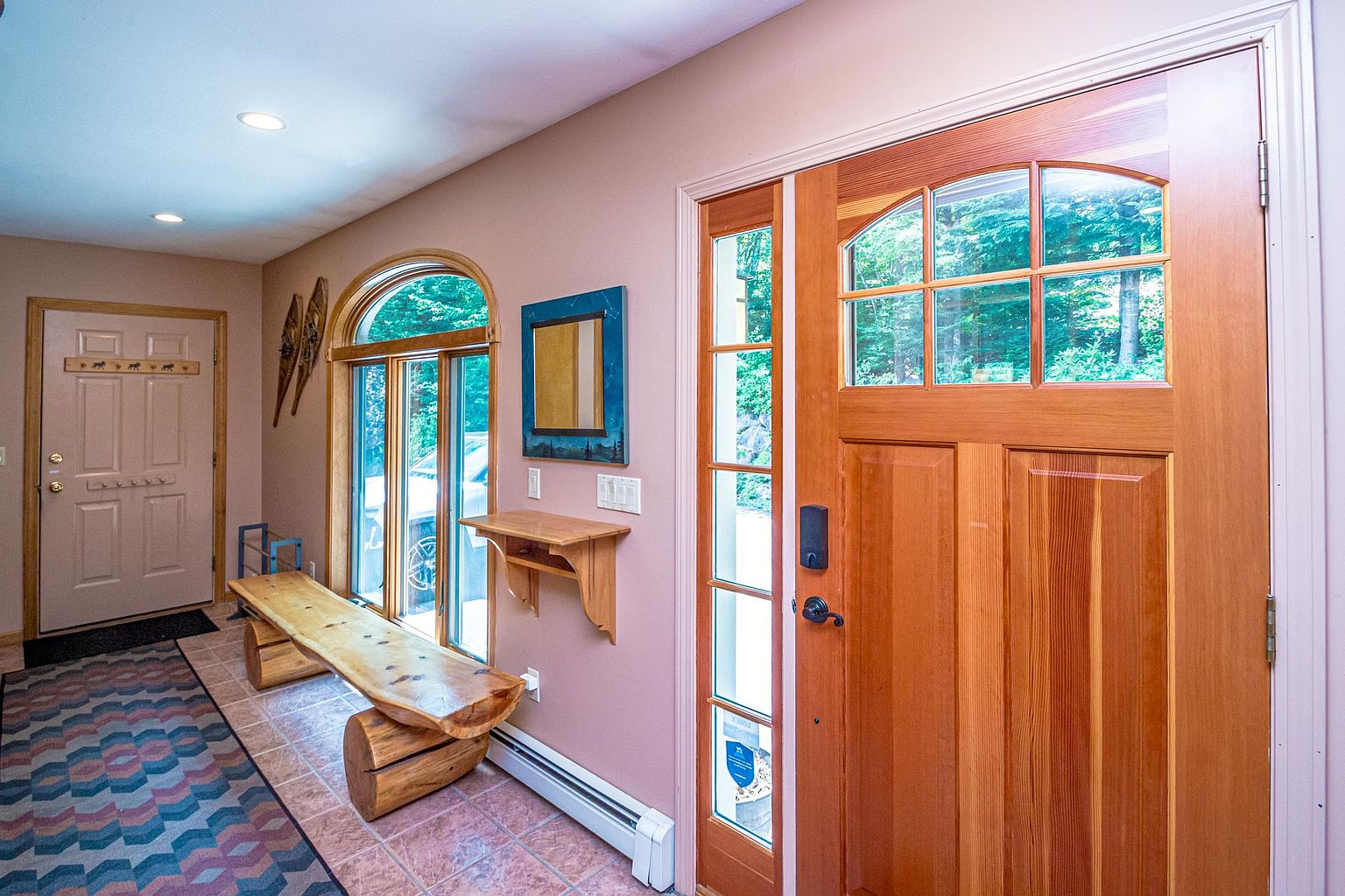 This inviting entryway features a warm, rustic aesthetic with a natural wood front door and a matching log-style bench. The space is finished with light-toned walls, tiled flooring, and a patterned runner, creating a welcoming transition area. A small wall-mounted shelf and a decorative mirror add functional charm, while large windows allow natural light to brighten the foyer.