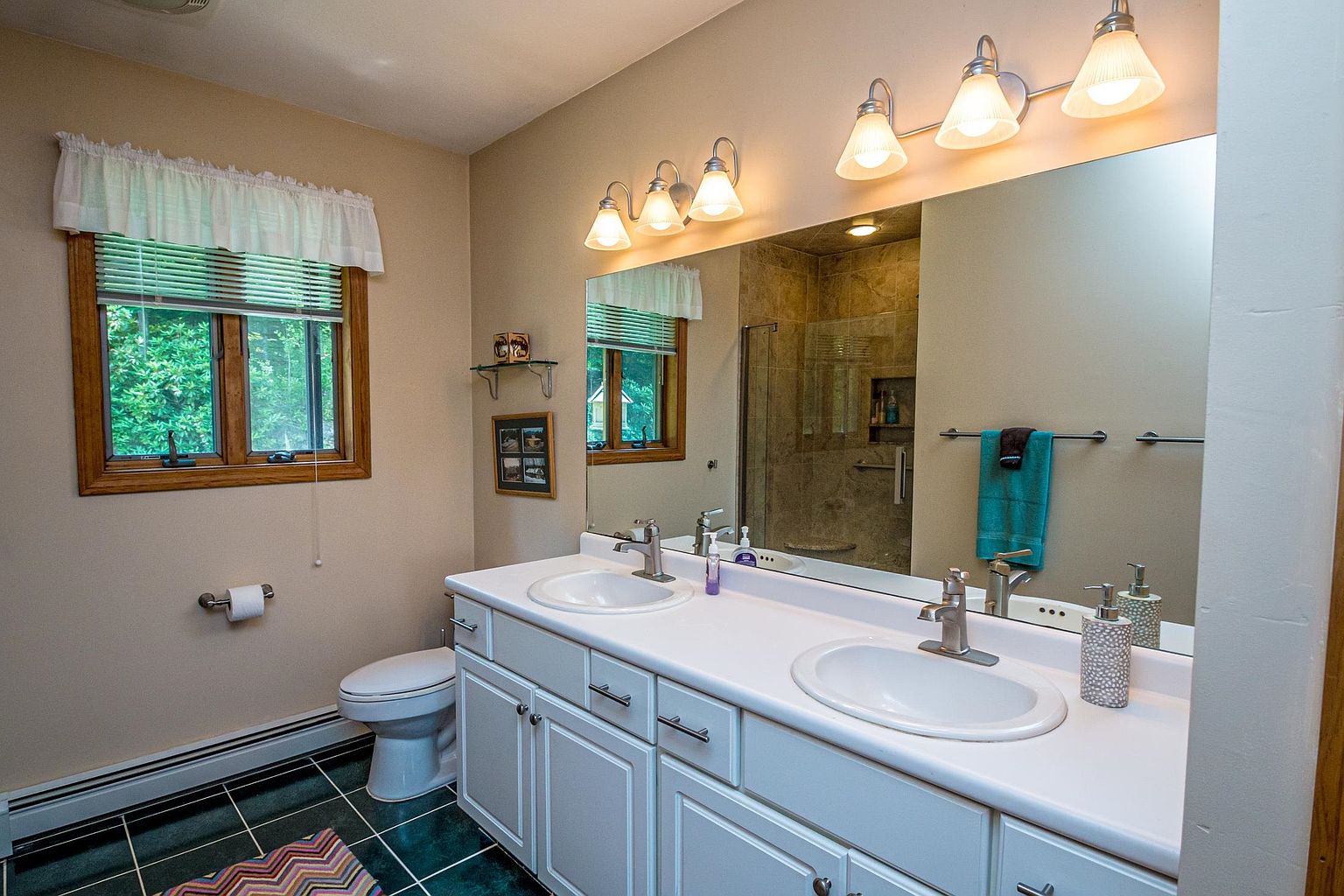 This bright, dual-vanity bathroom features white cabinetry topped with a solid-surface counter and two integrated sinks. A large, wide mirror spans the wall above the vanity, reflecting the walk-in shower with glass doors and tile surround, while a window with a valance provides natural light. The room is finished with dark, patterned floor tiles and a neutral wall color, creating a clean and functional space.